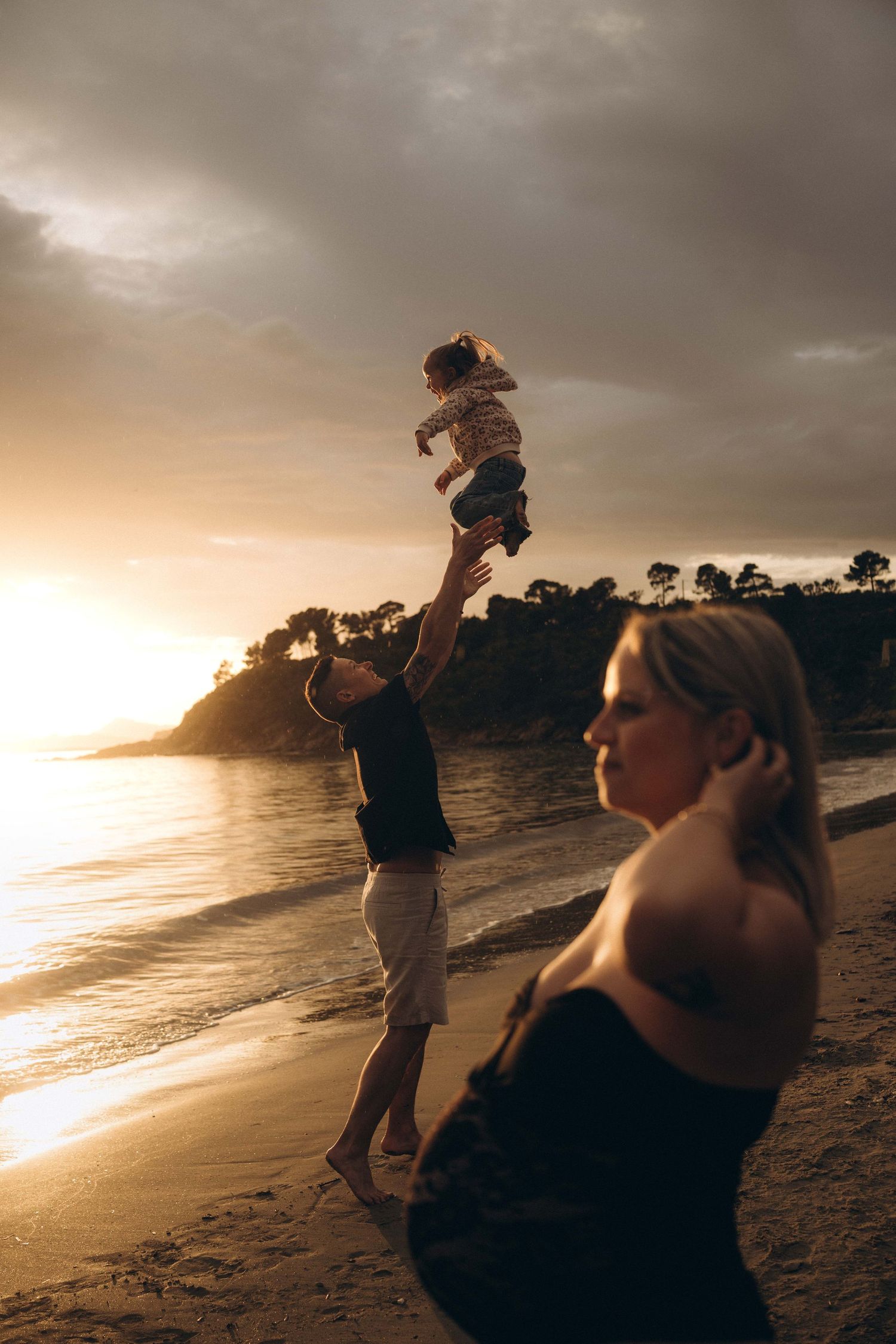 Family enjoying a beach sunset, father tossing child in the air while mother watches, golden hour sky backdrop.