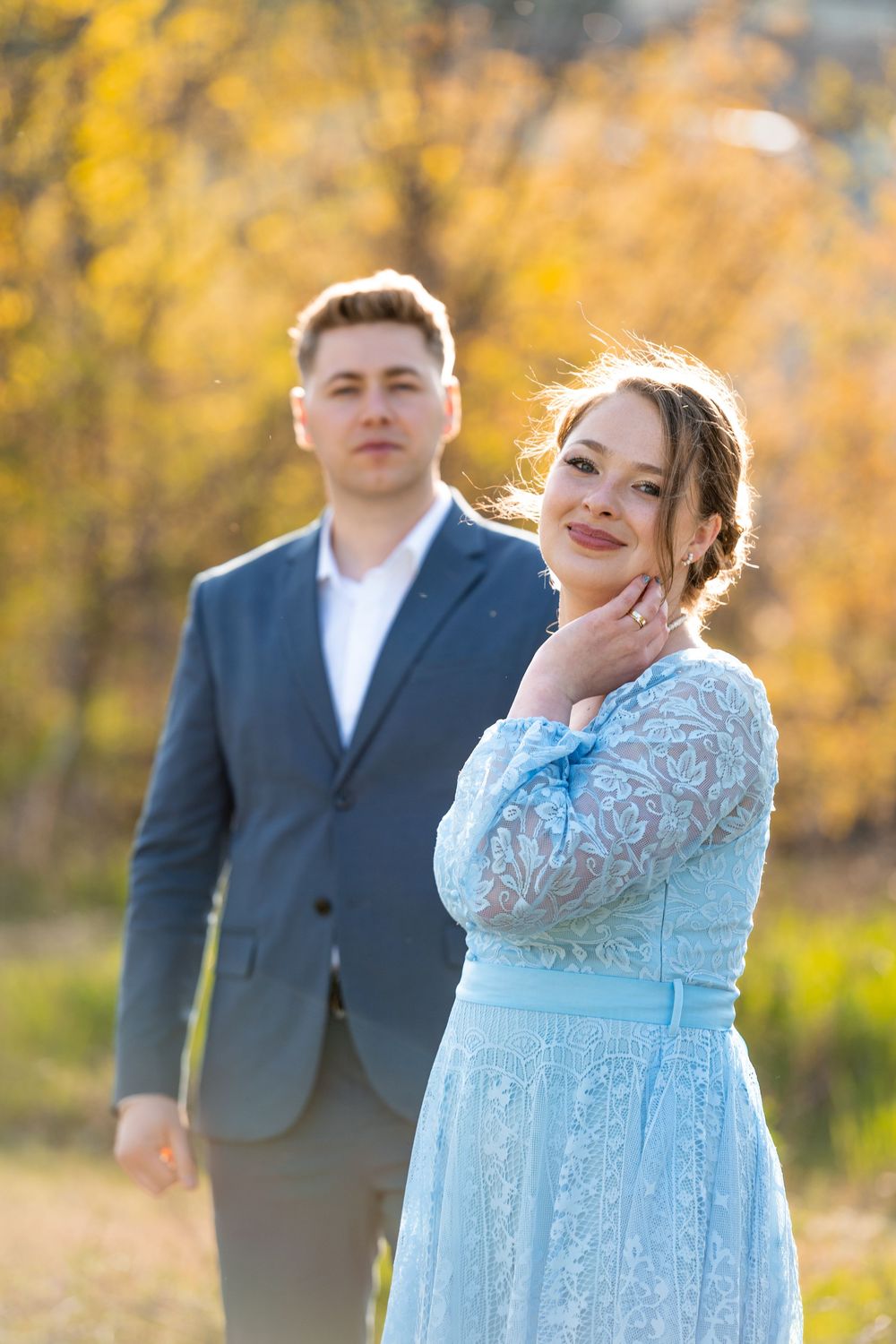 A smiling woman in a blue lace dress poses with a man in a suit amid golden autumn foliage.