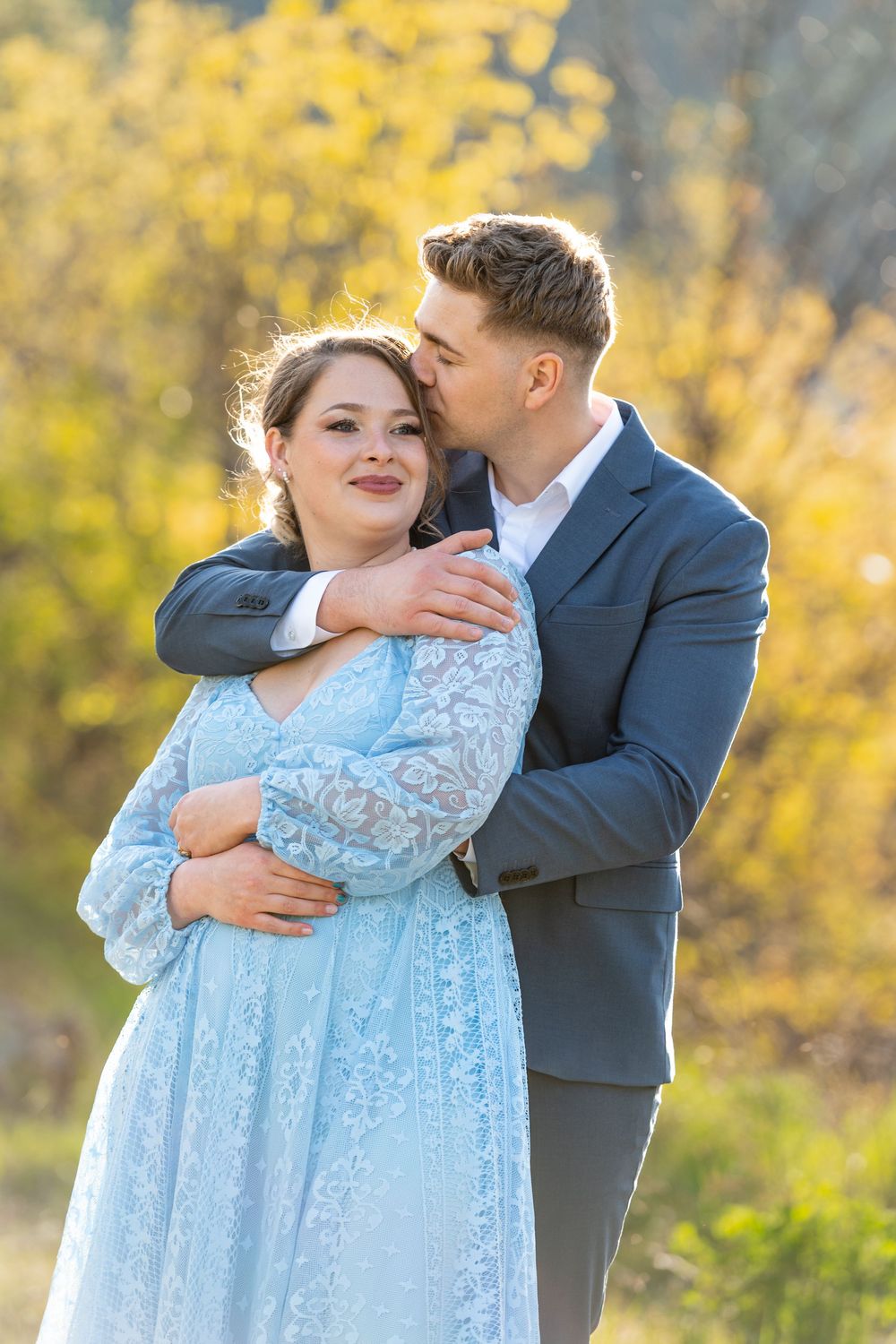 A couple embraces outdoors in autumn, woman in blue lace dress, man in suit jacket kissing her cheek.
