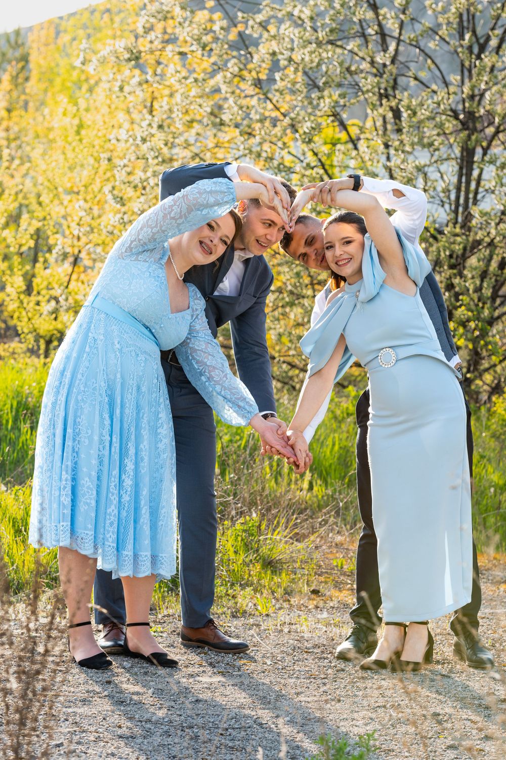 Four people in blue formal wear form a circle with hands joined outdoors near blooming trees in spring.