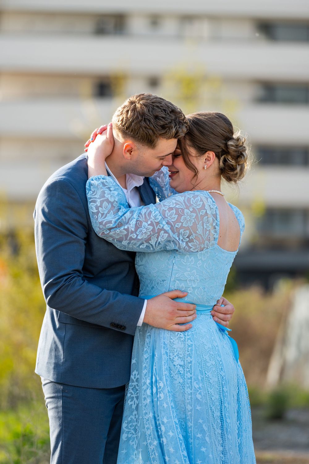 Couple in formal wear embracing outdoors, man in navy suit and woman in blue lace dress touching foreheads romantically.