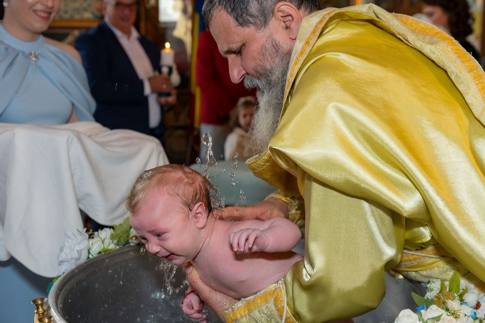 Priest in golden vestments performing Orthodox Christian infant baptism, immersing baby in baptismal font.