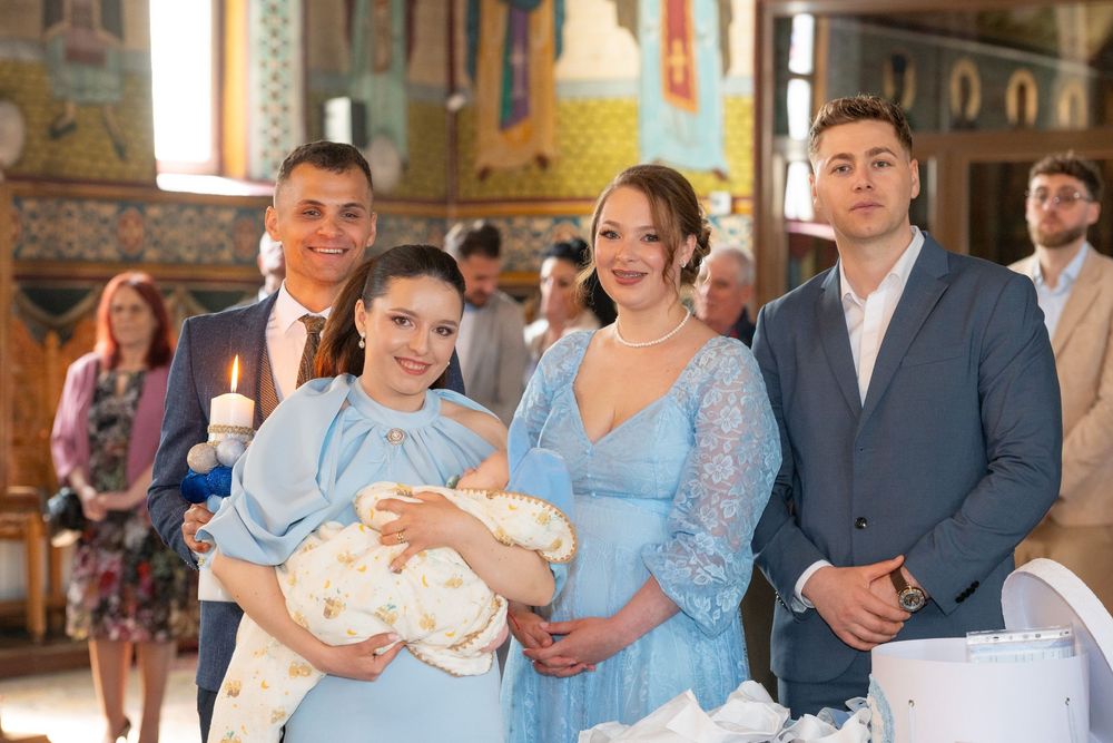 Family gathered in a church for a baptism ceremony, with parents holding a baby in white christening outfit.