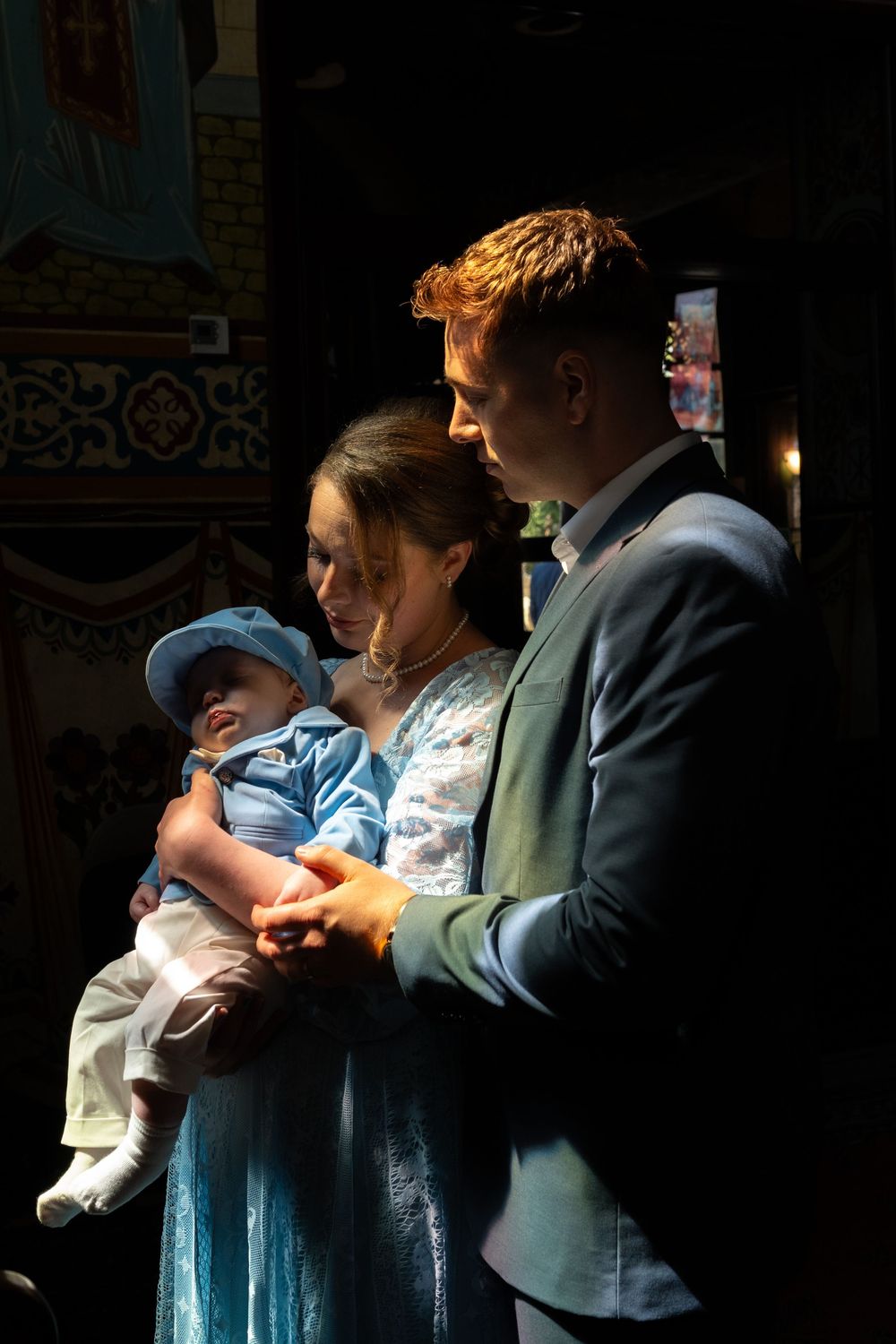 Young couple in formal attire holding a baby in a blue outfit inside a decorated church.