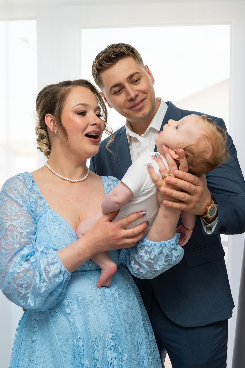 Young couple in formal attire lovingly holding their baby, woman in blue lace dress with pearl necklace, man in navy suit.