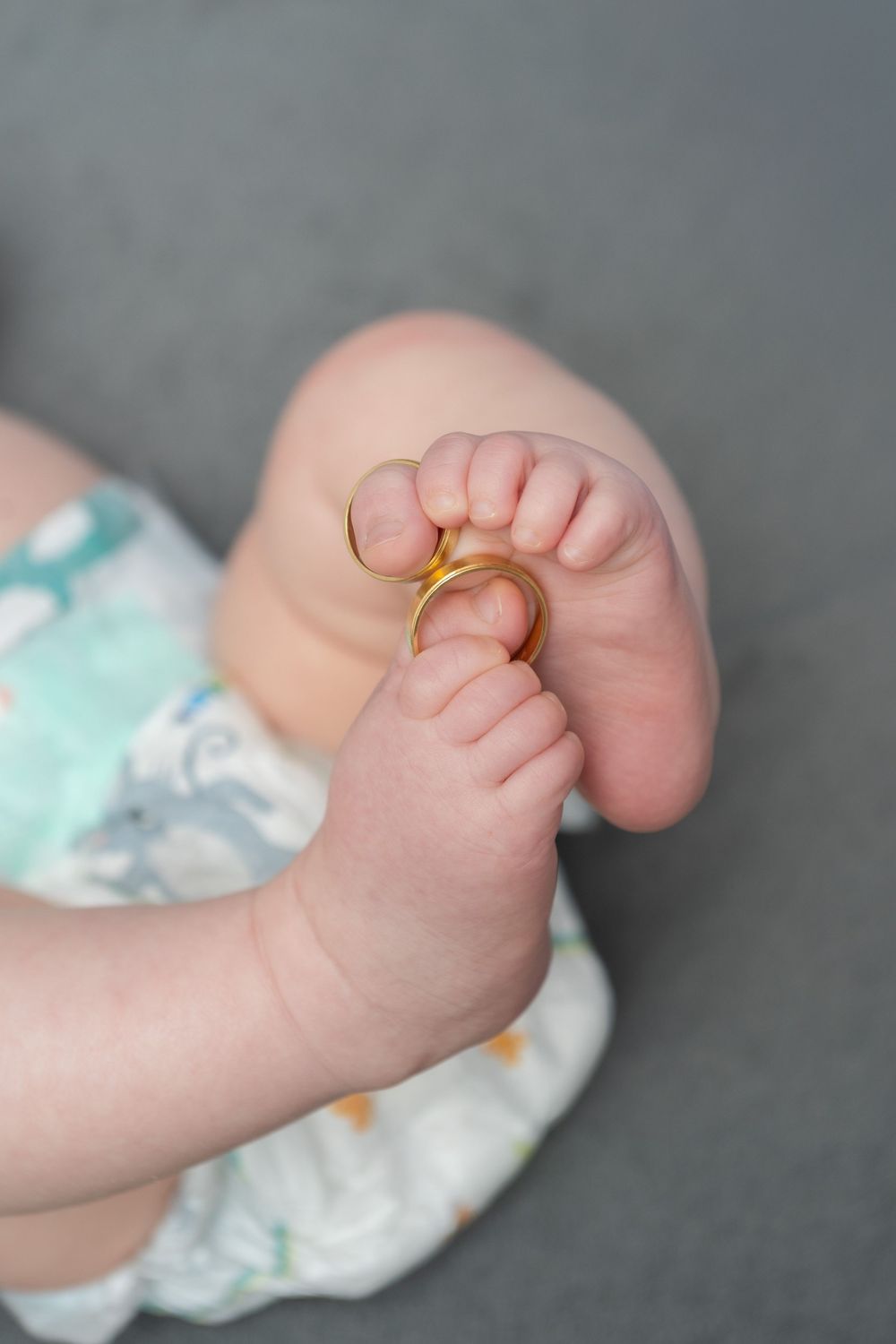 Close-up of chubby baby feet holding a gold wedding ring, wearing a floral onesie on gray background.