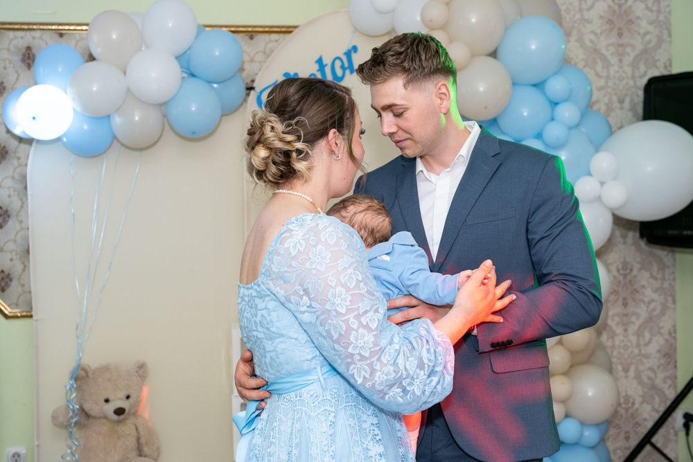 Couple in formal attire tenderly holding a newborn baby at a blue-themed baby shower with balloon arch.
