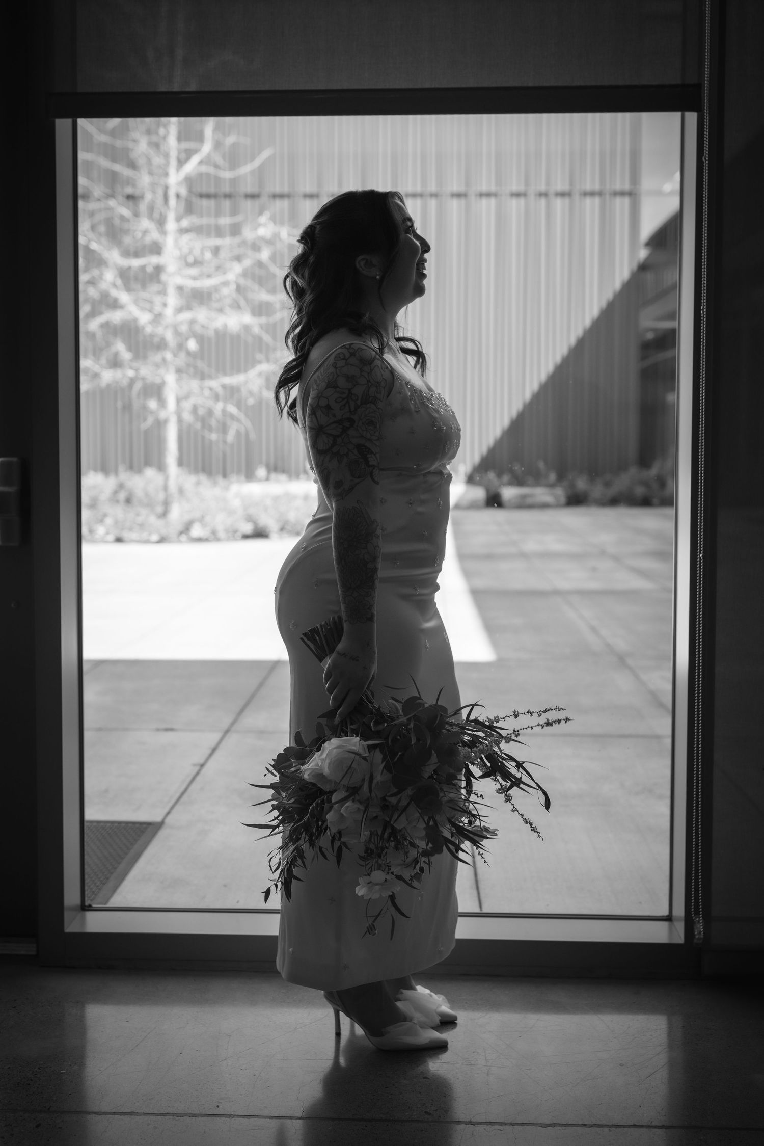 Silhouetted bride in wedding gown holding bouquet stands before large glass door in black and white.