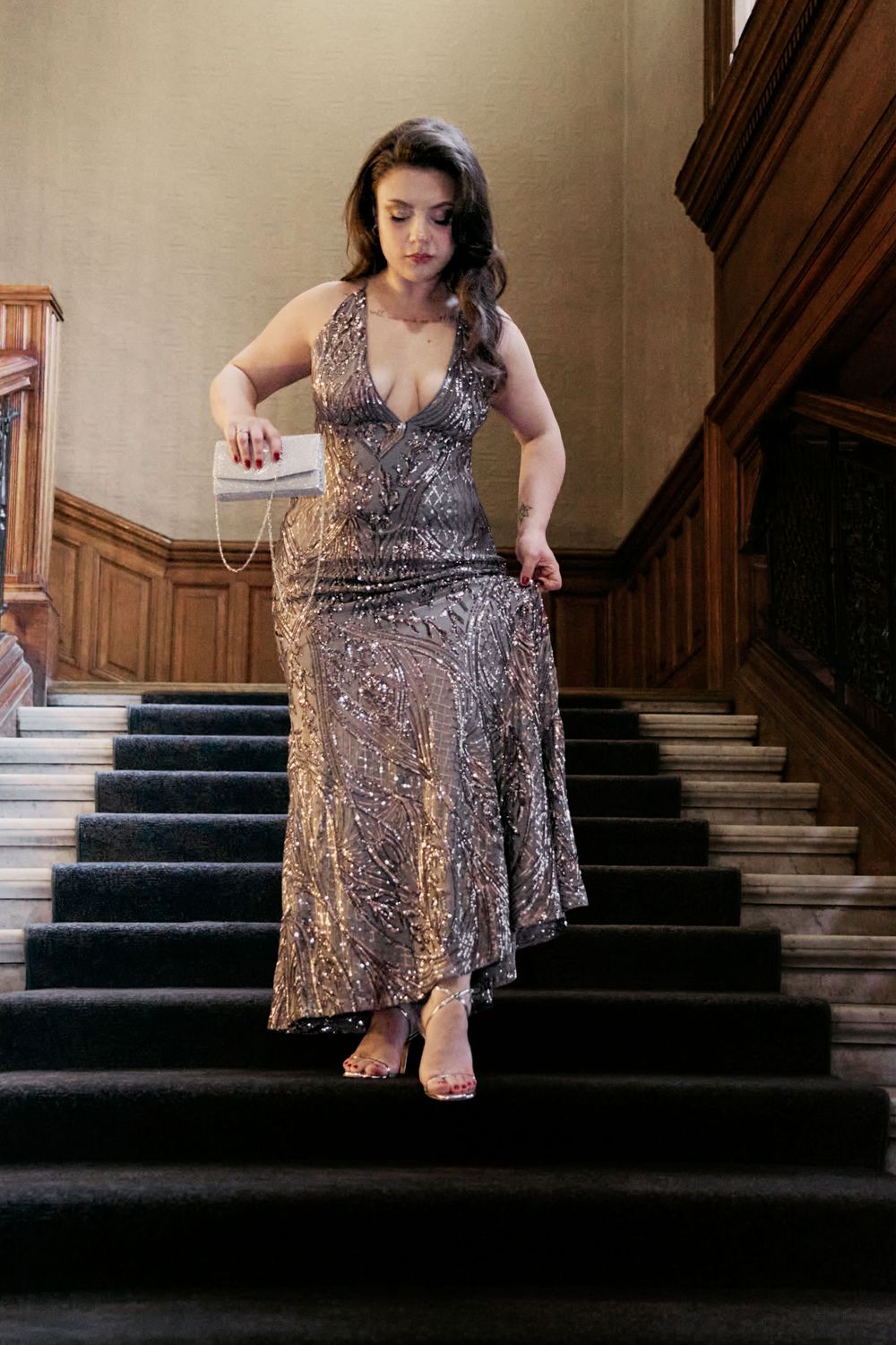 Woman in silver sequin gown descending grand staircase, holding clutch purse, elegant formal attire.