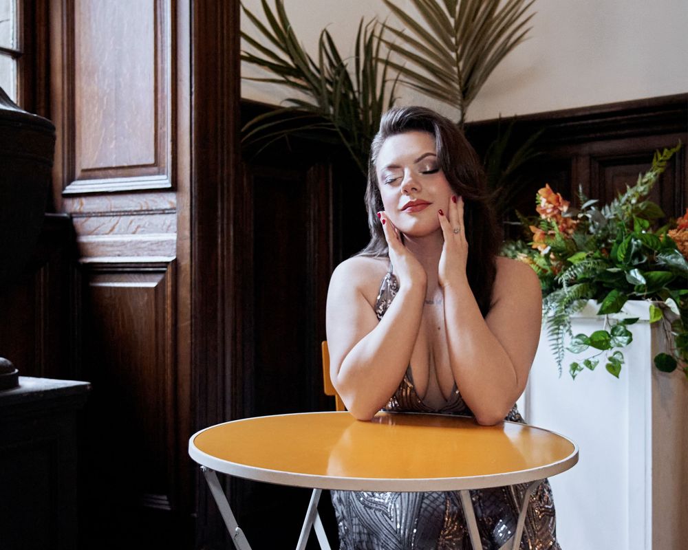 A woman in a sequined dress poses gracefully at a yellow round table beside tropical flowers in an elegant wood-paneled room.