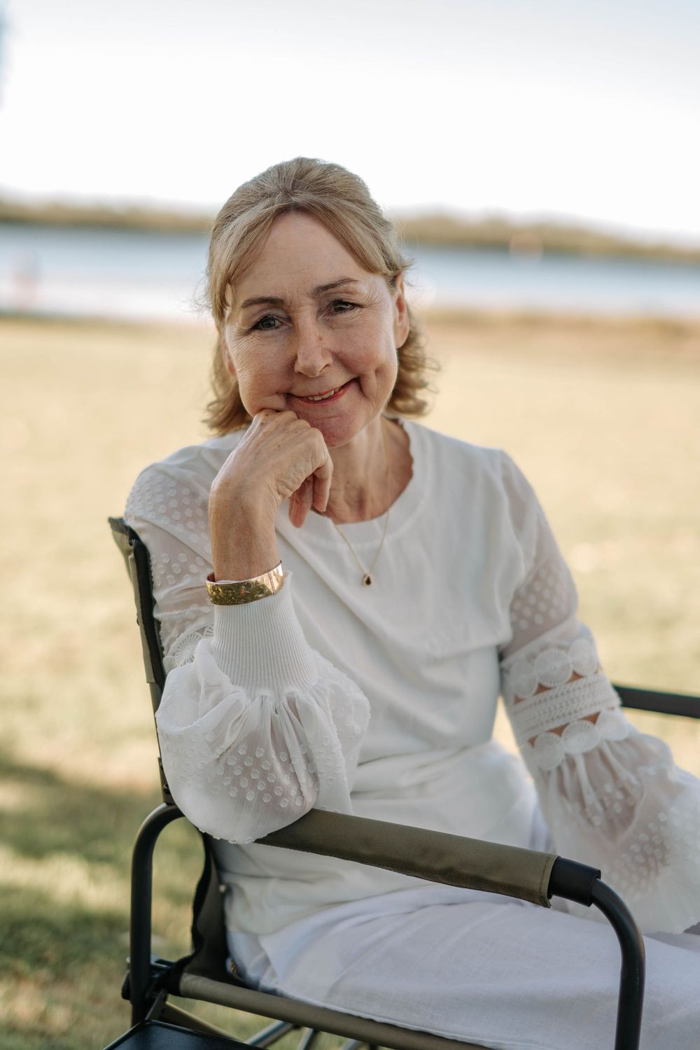 Smiling middle-aged woman in white dress sitting outdoors by a lake, resting chin on hand.