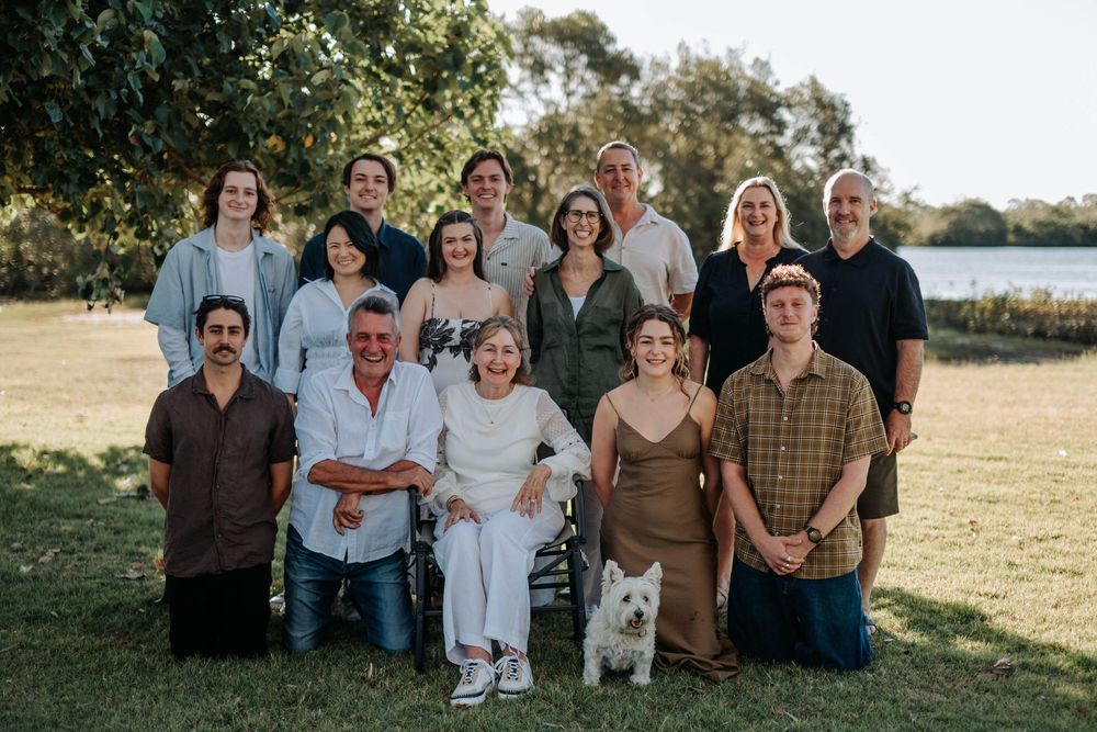 Large family group portrait outdoors on green grass near a lake, including a white dog, smiling together on a sunny day.