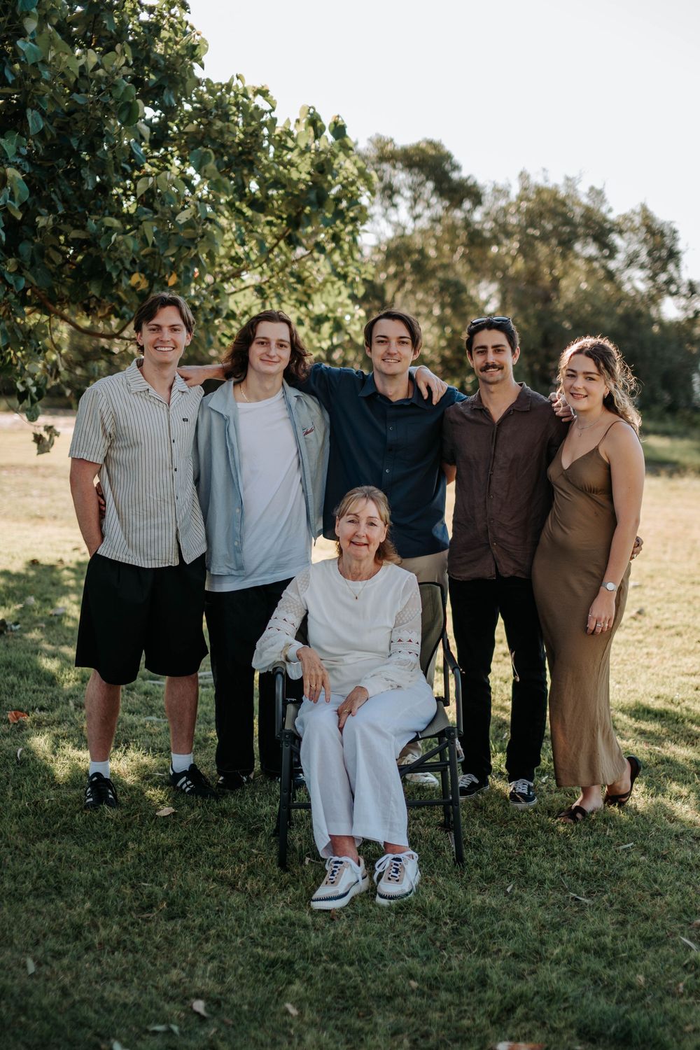 Family of six posing outdoors with elderly woman in wheelchair, smiling together under green trees on grass.