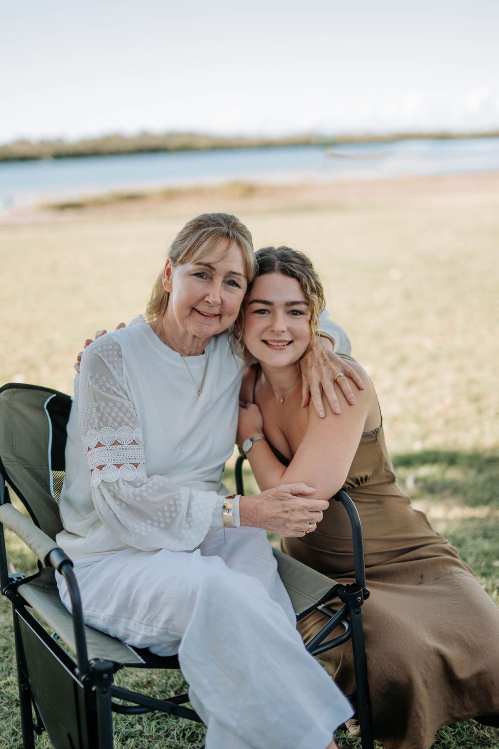 Two women smile and embrace outdoors near a waterfront, one seated in a camp chair wearing white, the other in a golden dress.