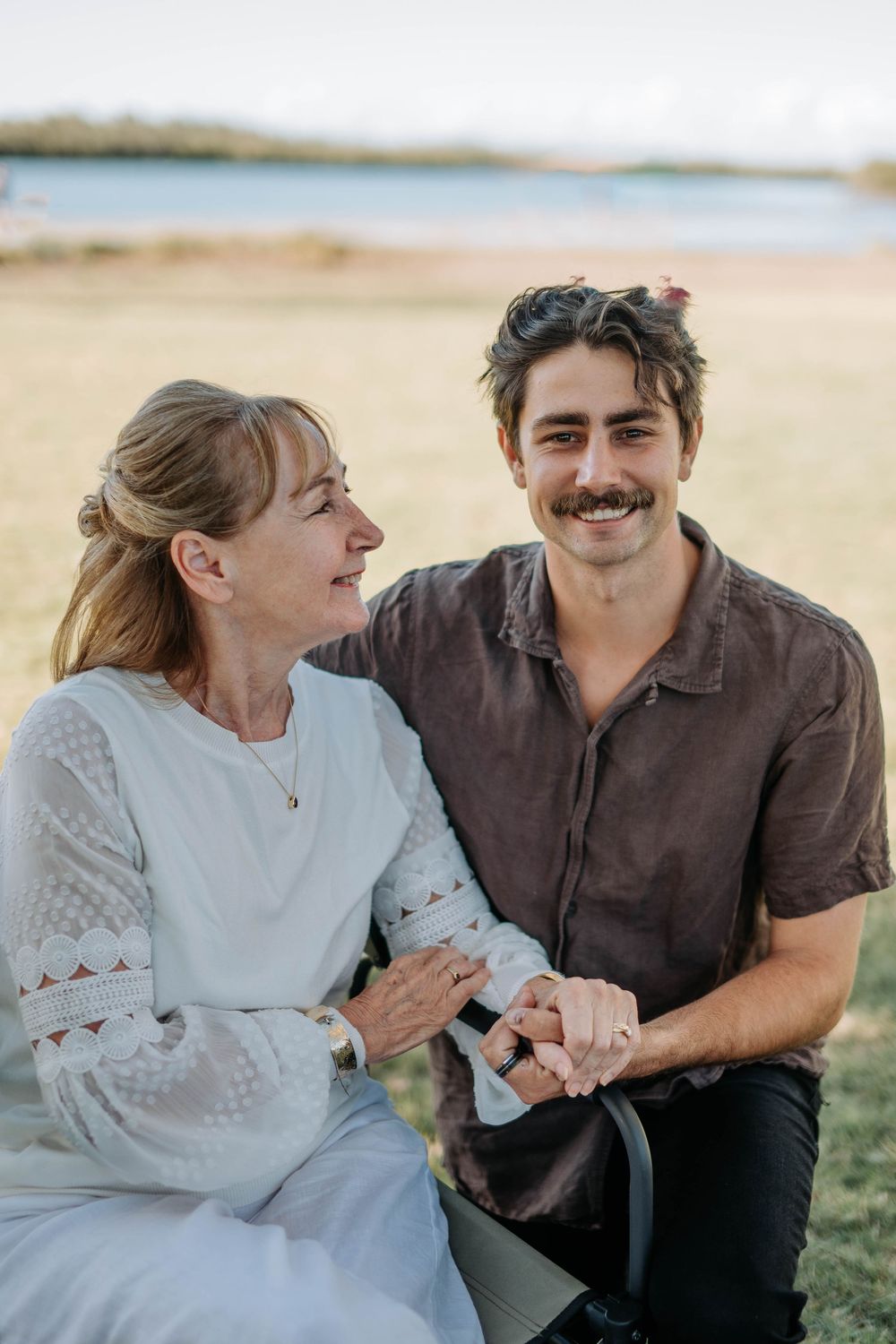 A smiling young man with mustache holds hands with an older woman outdoors near a lake.