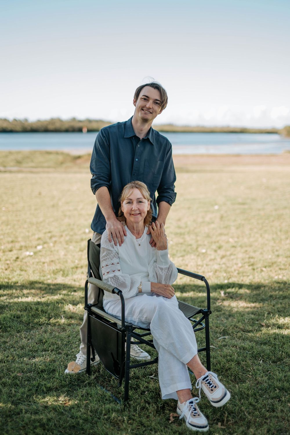 Young man stands behind elderly woman seated in chair outdoors near waterfront, both smiling warmly on sunny day.