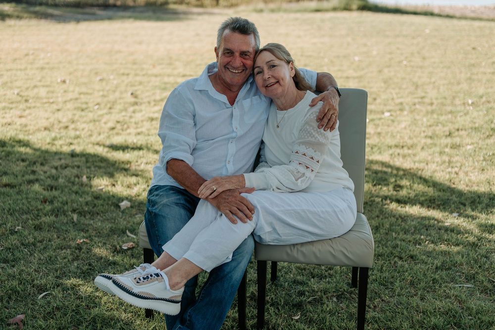 Smiling middle-aged couple sitting together on a chair outdoors on green grass, wearing casual white outfits.