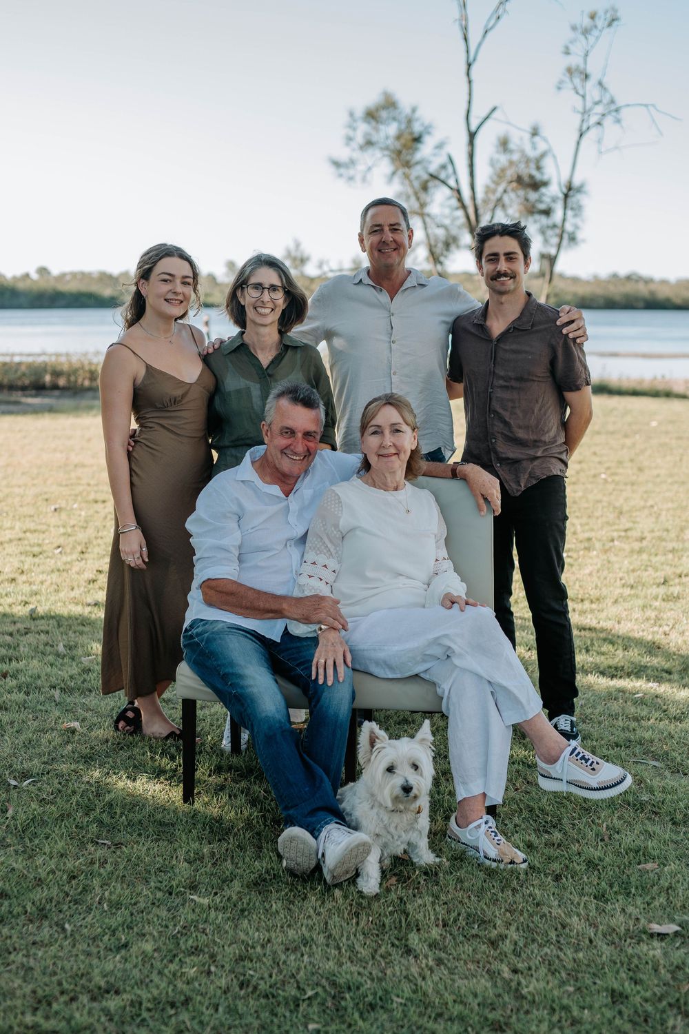Family of six poses outdoors near a lake with a white Westie dog on green grass at sunset.