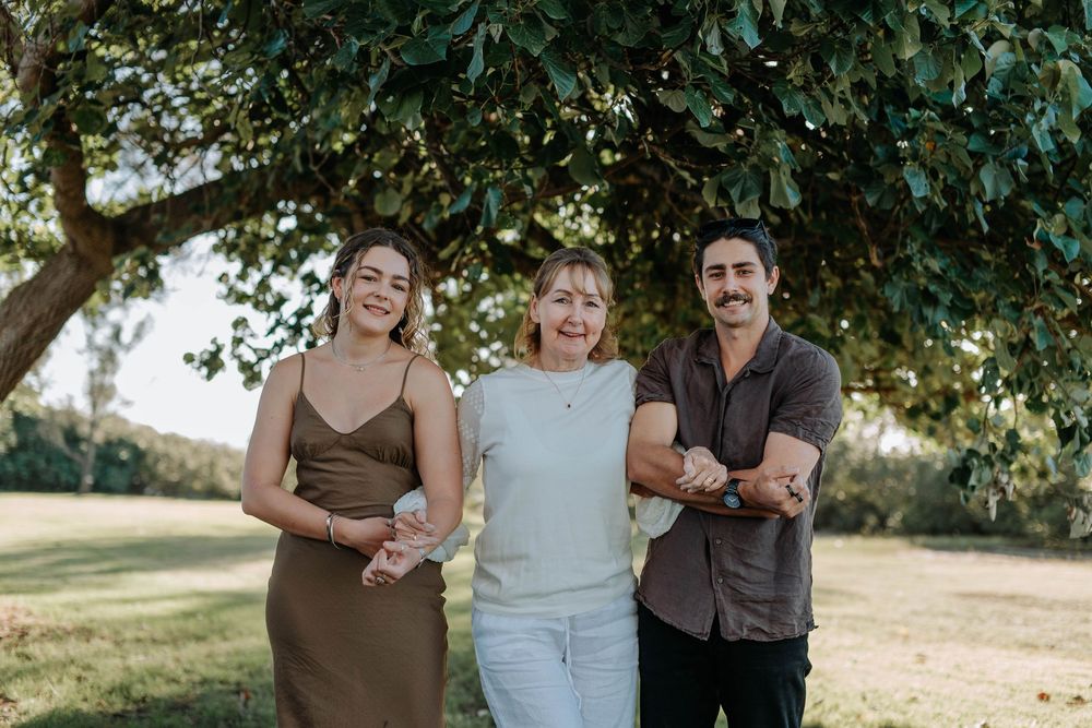 Three adults smiling and posing together under a large tree outdoors in natural light.