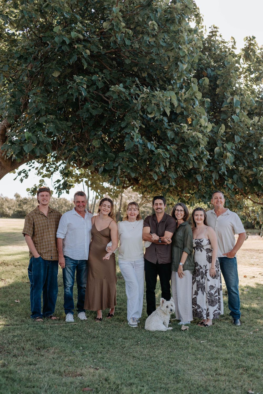 Extended family group of eight adults with white dog poses under large tree in park setting.