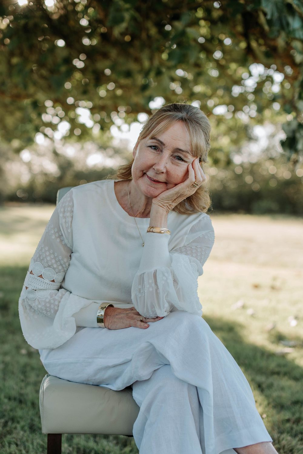 Smiling older woman in white dress sitting outdoors on chair, resting cheek on hand, with green trees in background.