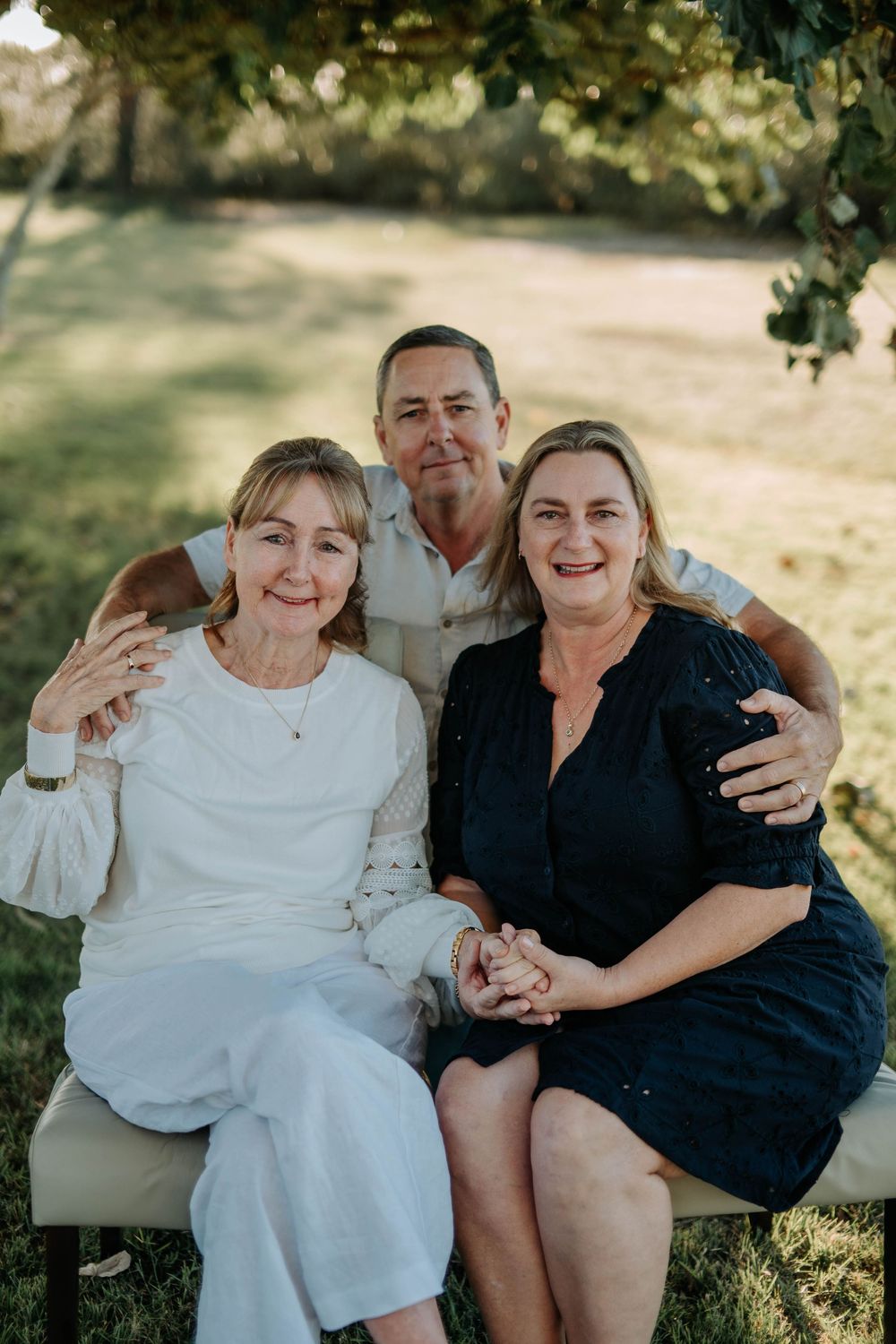 Three smiling family members pose together outdoors on a bench surrounded by lush green trees.