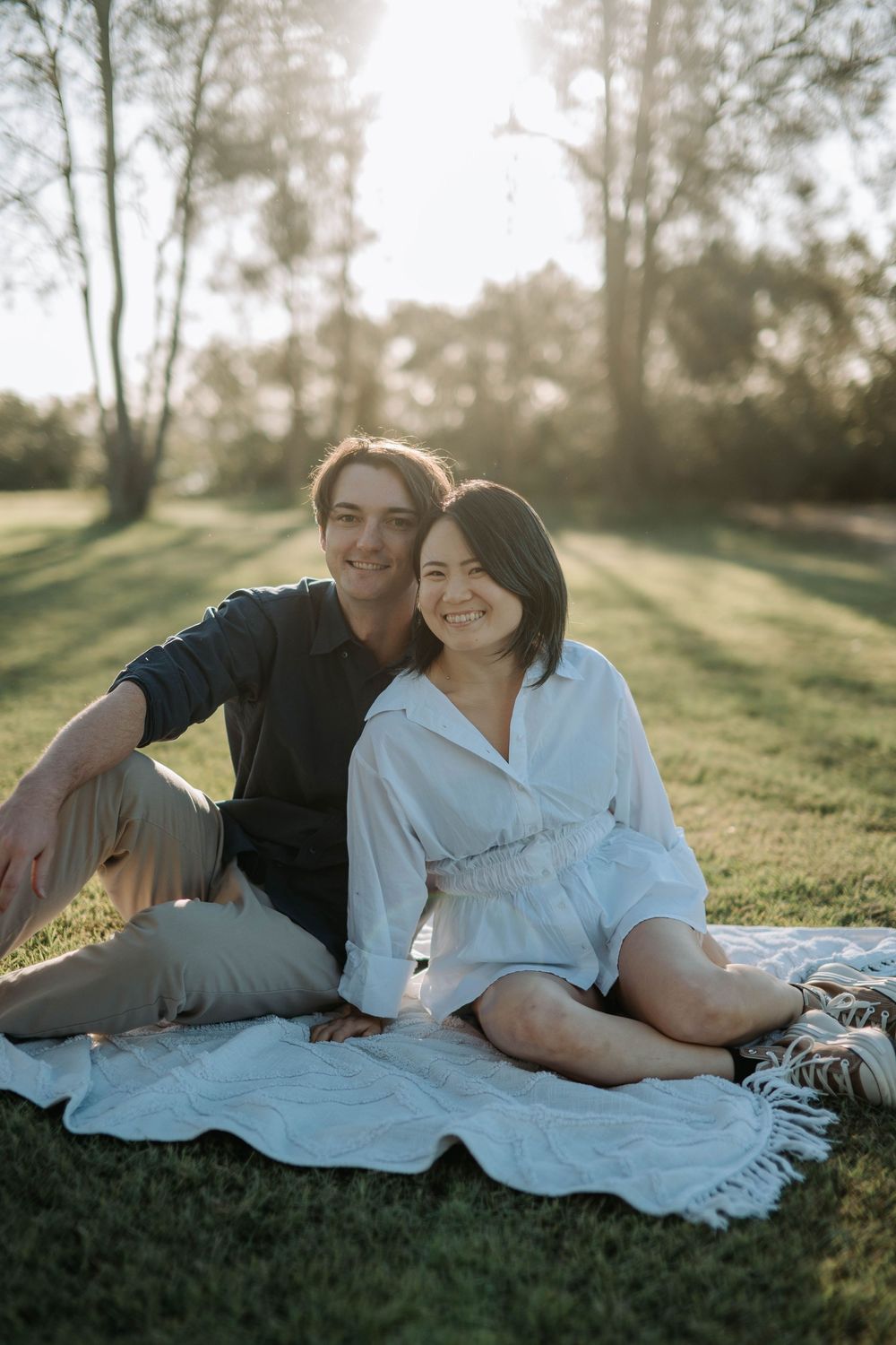 Young couple sits together on white blanket in sunlit park, smiling at camera during golden hour.
