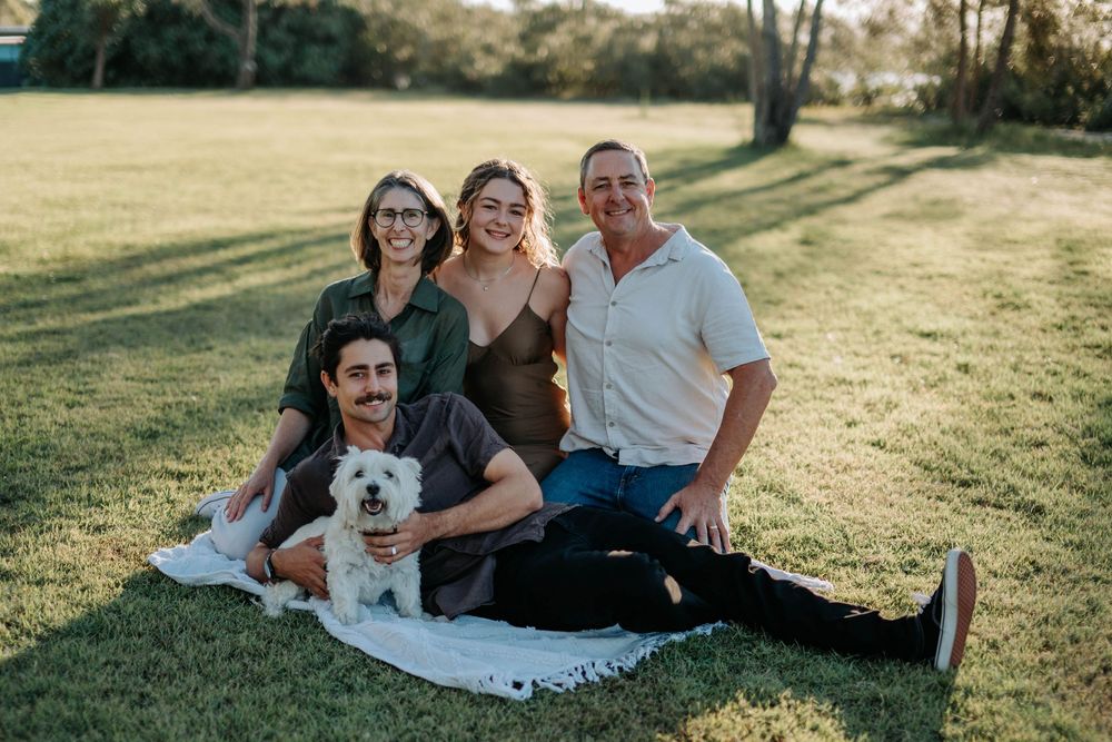 Family of four smiling outdoors on a blanket in a sunny park with a small white fluffy dog.
