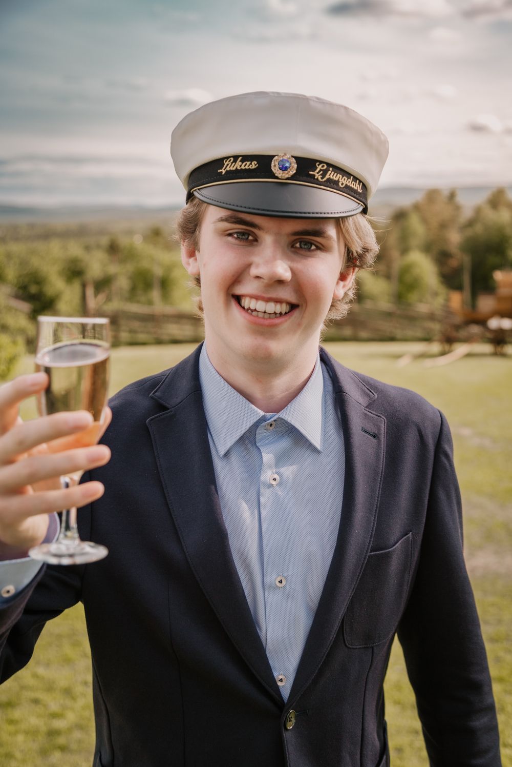 Young man in navy suit and graduation cap smiling while raising a champagne glass outdoors.