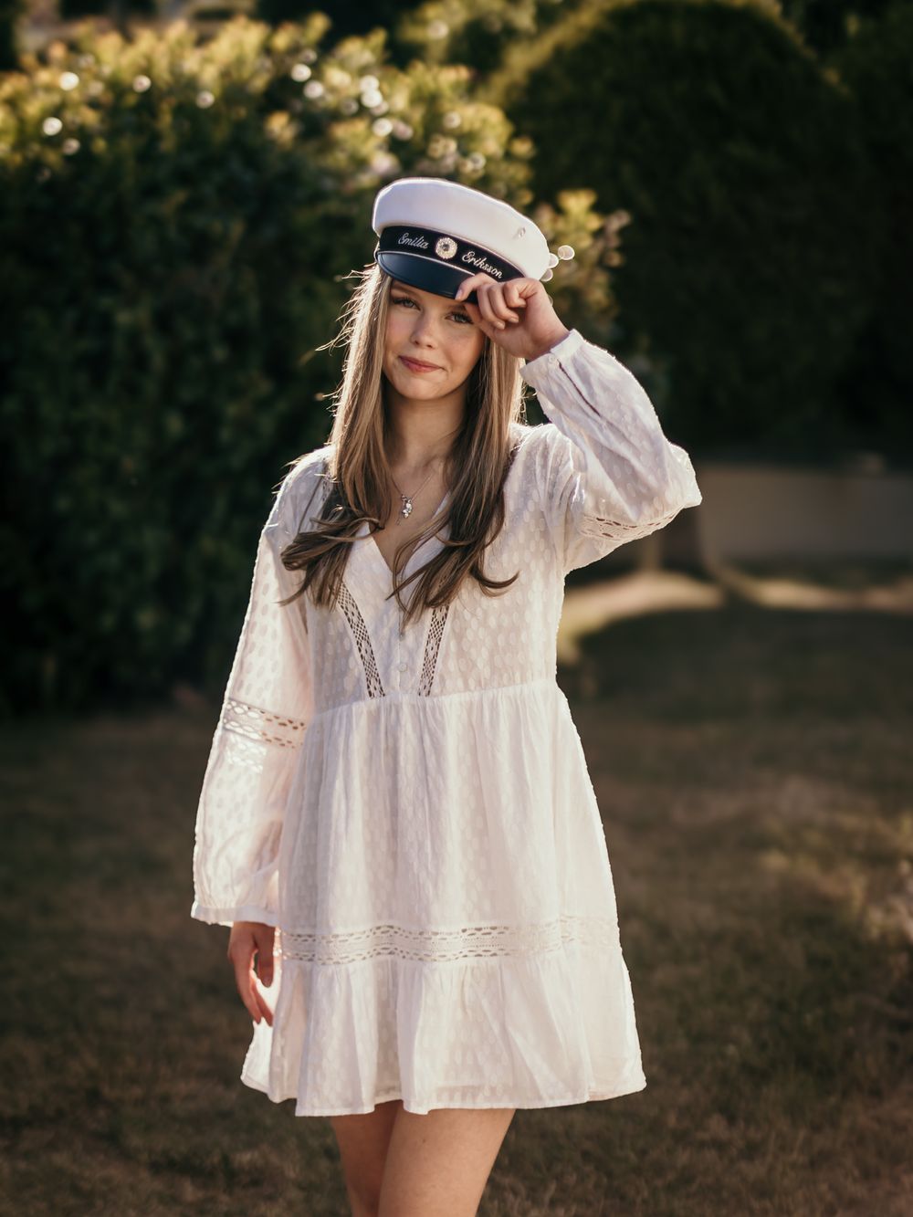 Young woman in white lace dress and navy captain hat poses outdoors near green bushes in warm sunlight.