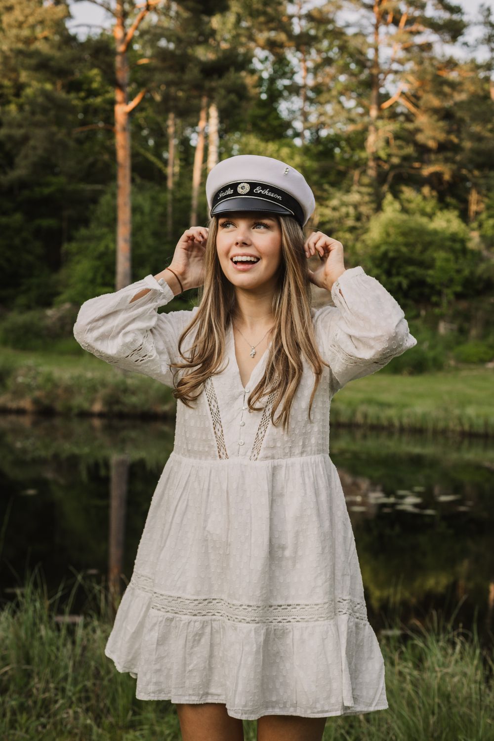 Young woman in white dress and sailor cap smiling by a forest pond.