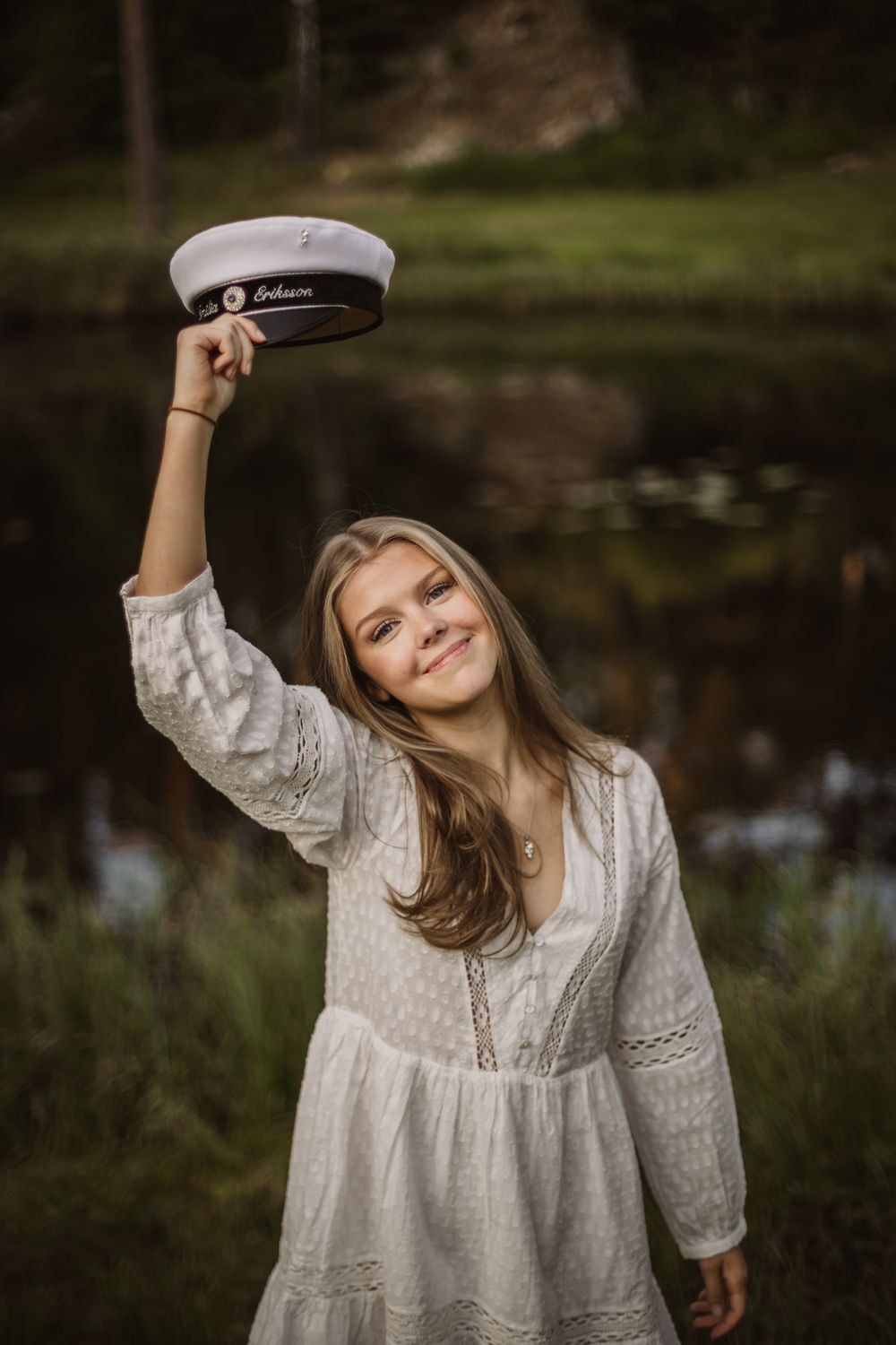 Young woman in white lace dress smiling while raising a white graduation cap outdoors near a pond.
