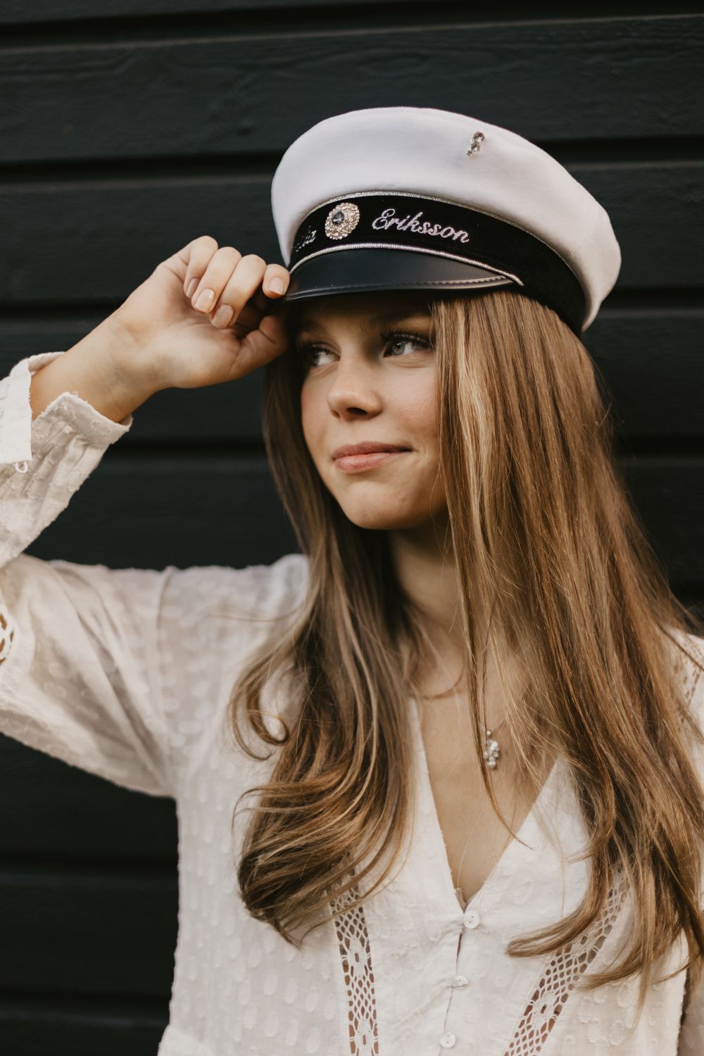 Young woman with long brown hair wearing a white sailor cap and lace blouse, posing against a dark wall.