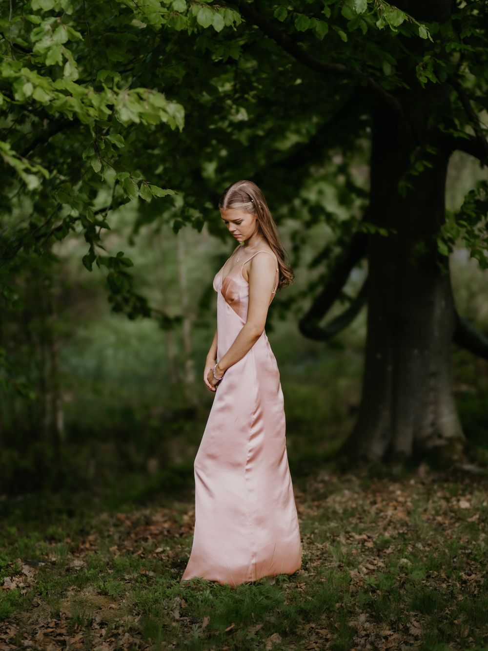 A young woman in a flowing pink satin gown stands gracefully beneath lush green trees in a serene forest setting.