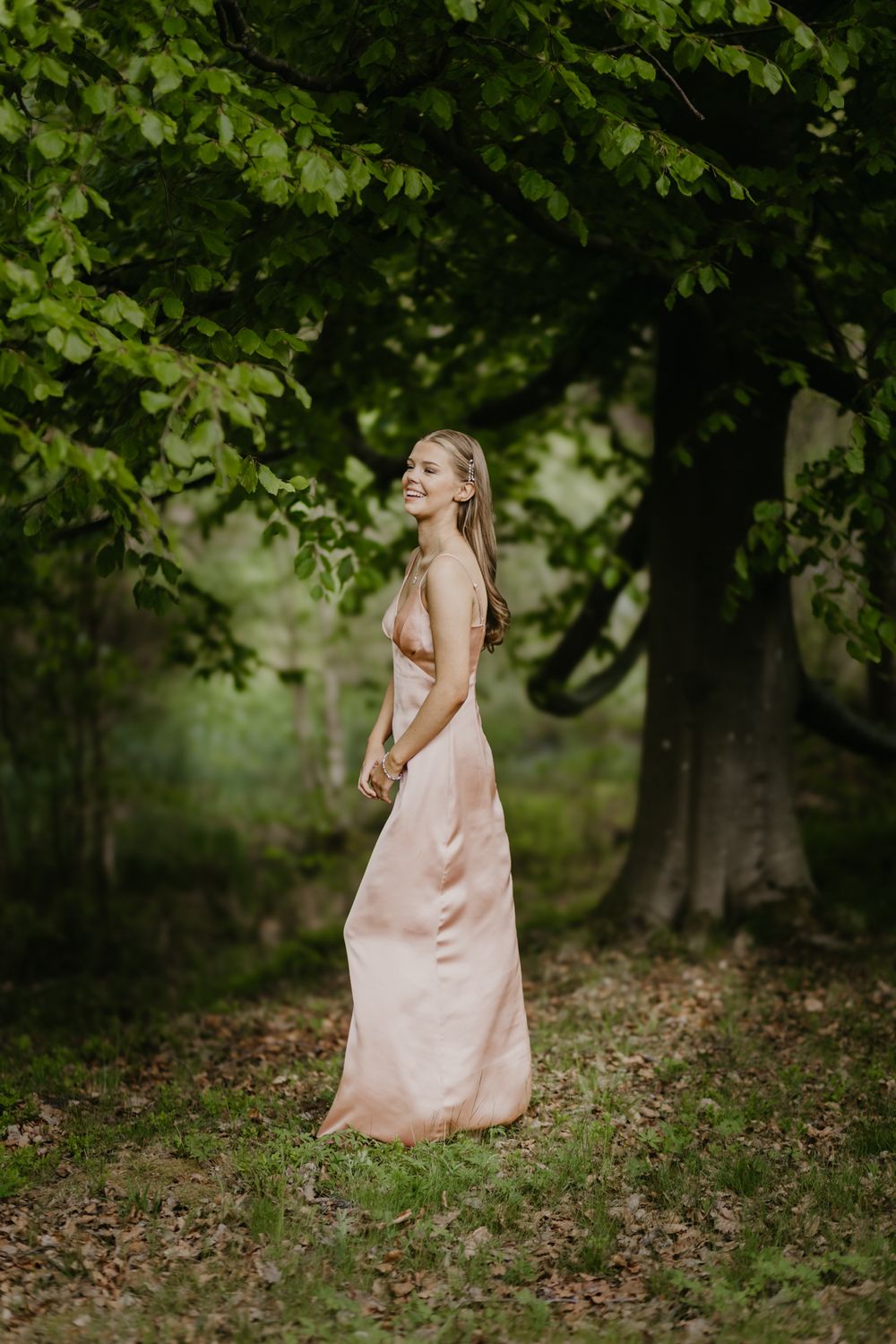 Smiling woman in elegant pink satin gown standing gracefully among lush green trees in a serene forest setting.