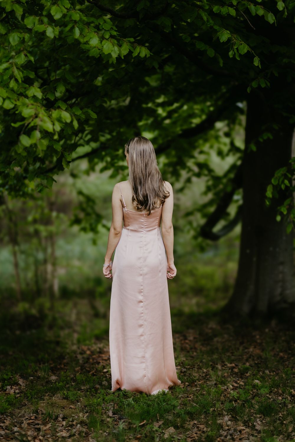 Woman in pink satin gown standing beneath lush green trees in a forest, viewed from behind.