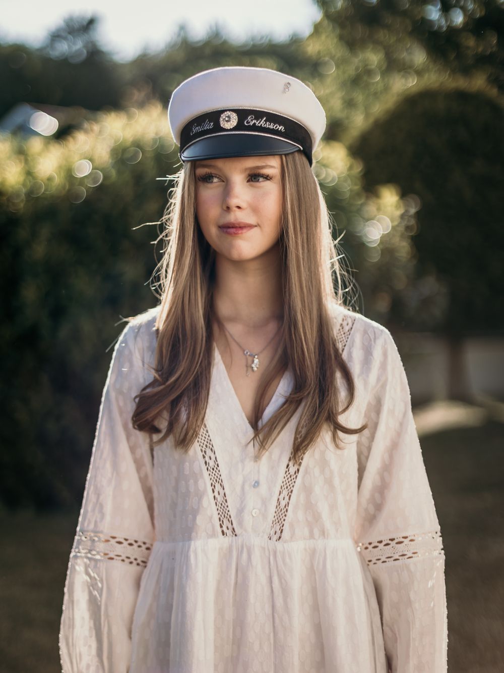 Young woman in white lace dress and personalized captain hat poses outdoors in warm golden light.