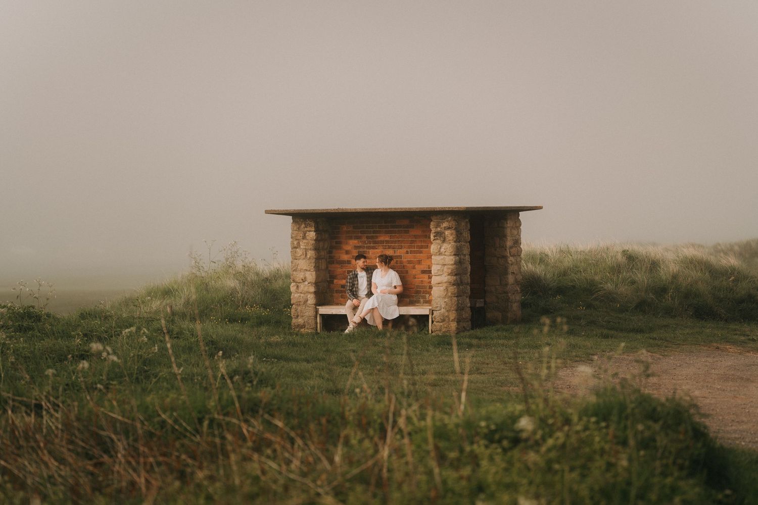 Couple sitting together in a rustic brick bus shelter surrounded by misty green fields.