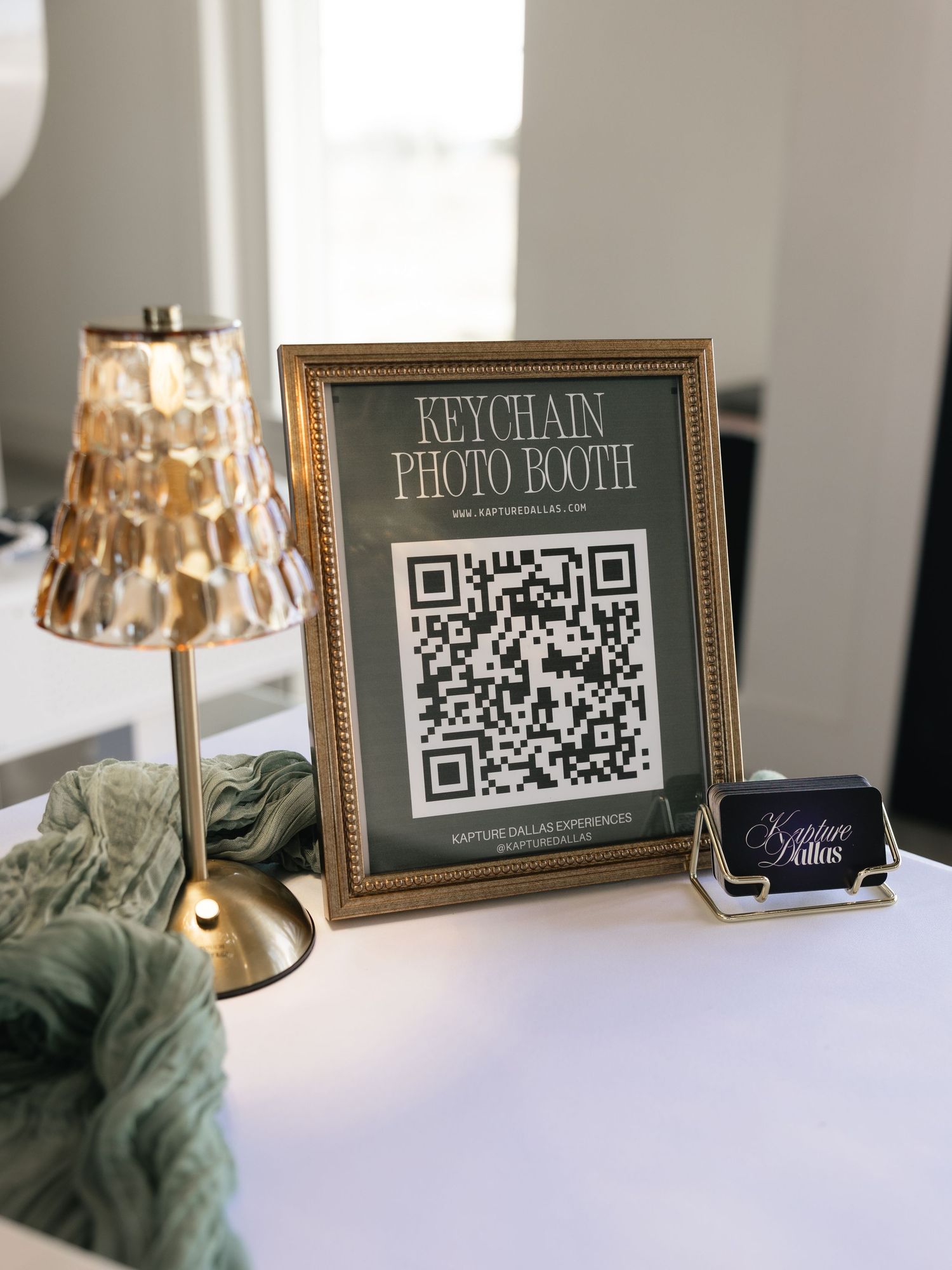 Gold-framed QR code sign for a Keychain Photo Booth display on a white table with a brass lamp and business cards.