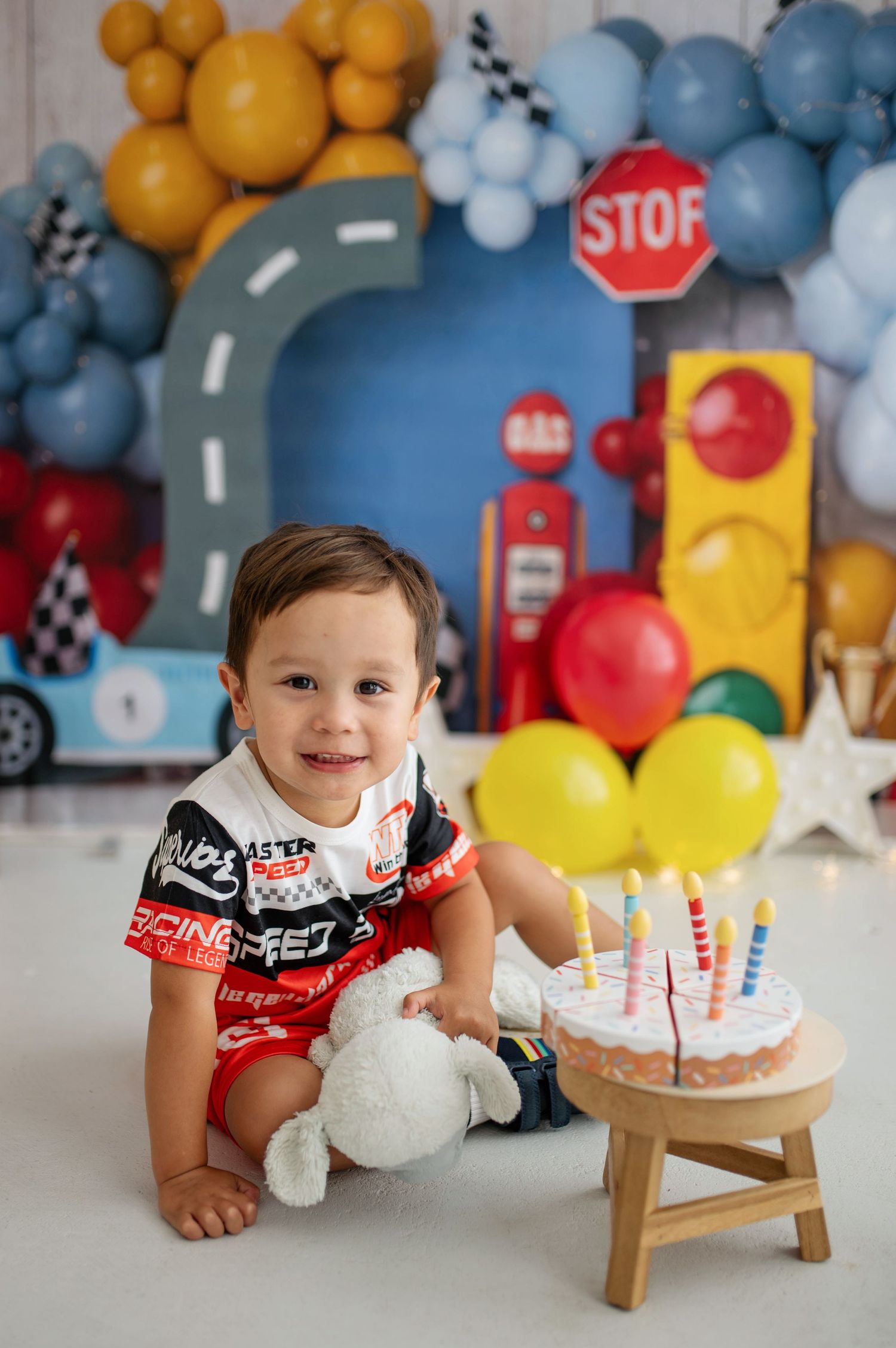 Toddler boy smiling at first birthday cake smash with race car theme, balloons, and stop sign backdrop.