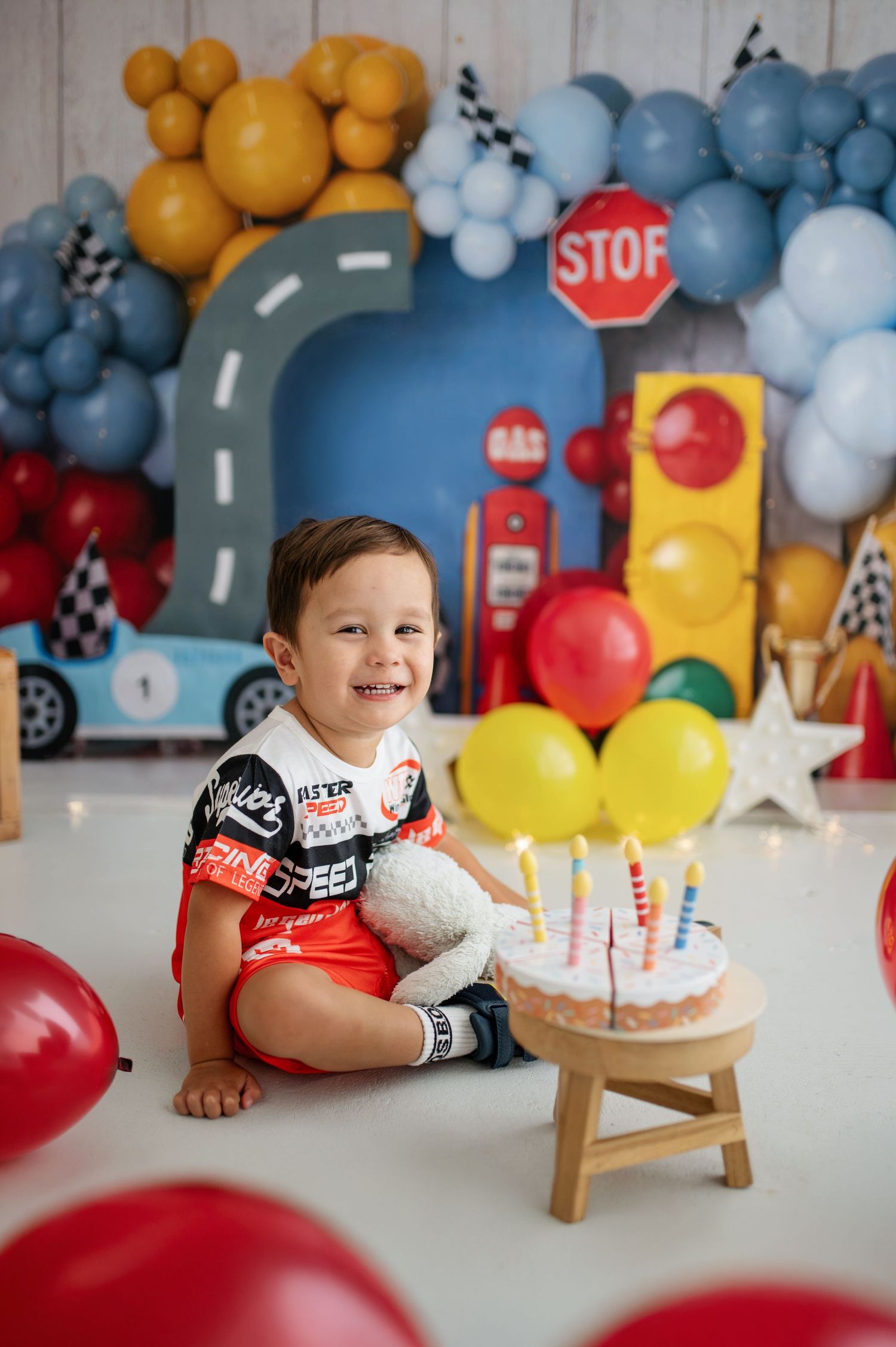 Smiling baby boy sitting beside a small birthday cake with candles, surrounded by colorful balloons and a racing theme backdrop.