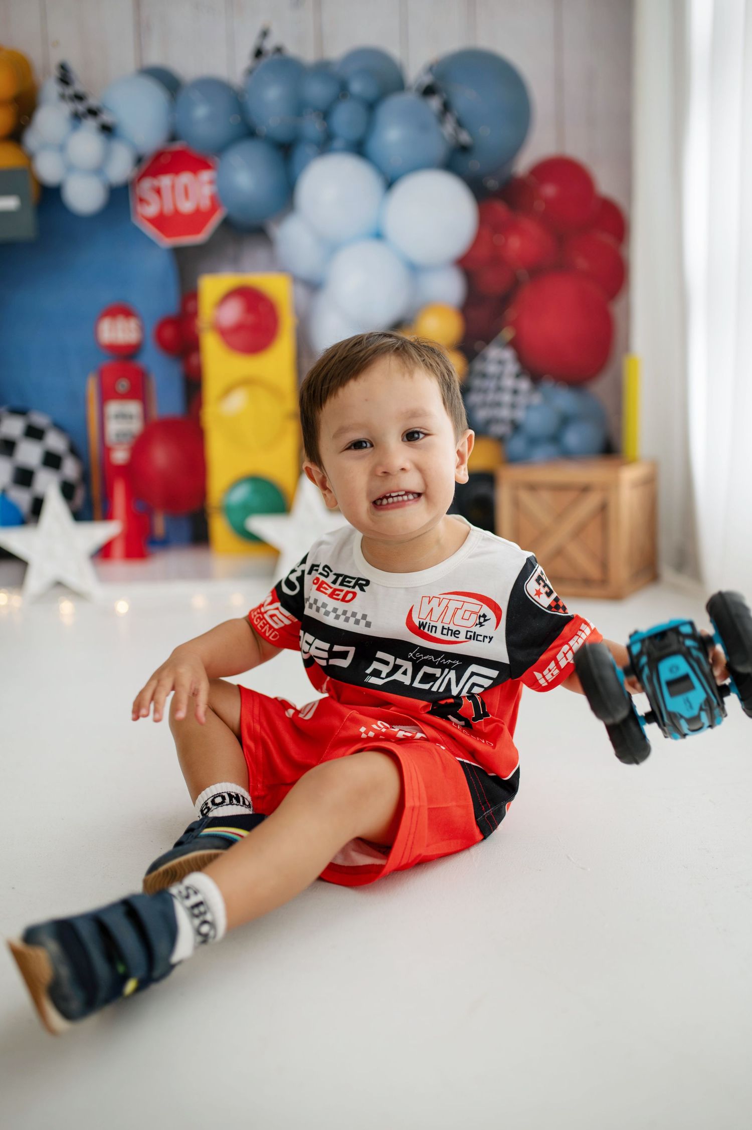 Toddler boy in racing outfit sits on white floor with toy car, balloon backdrop with stop sign decor.