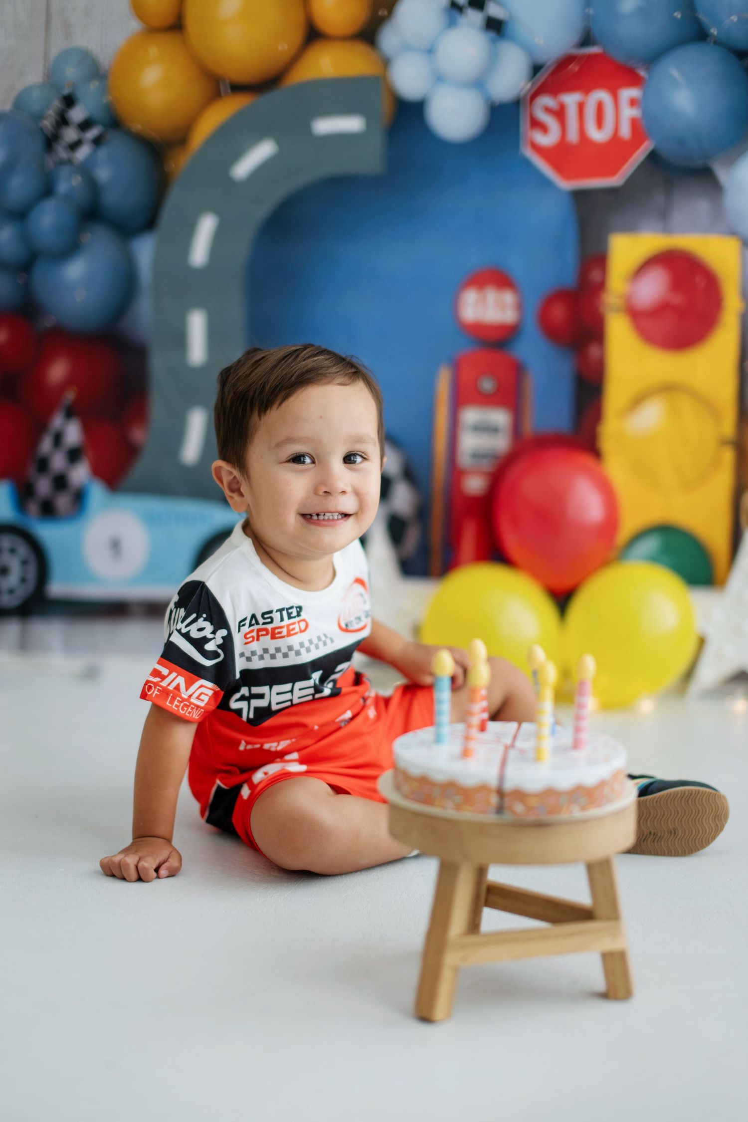 Smiling toddler boy in racing outfit sits beside birthday cake with candles, race car themed party backdrop.