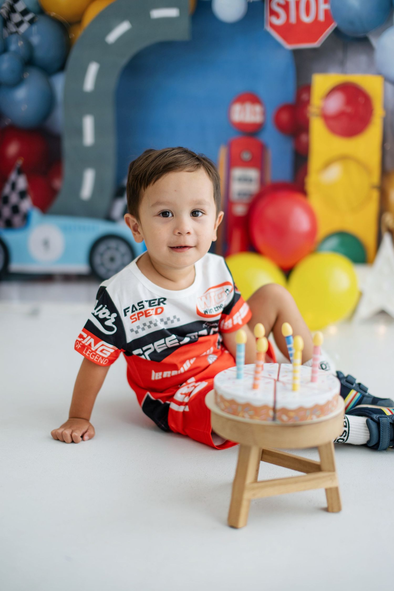 Toddler boy in racing outfit sits beside birthday cake with candles, surrounded by colorful race car themed party decorations.
