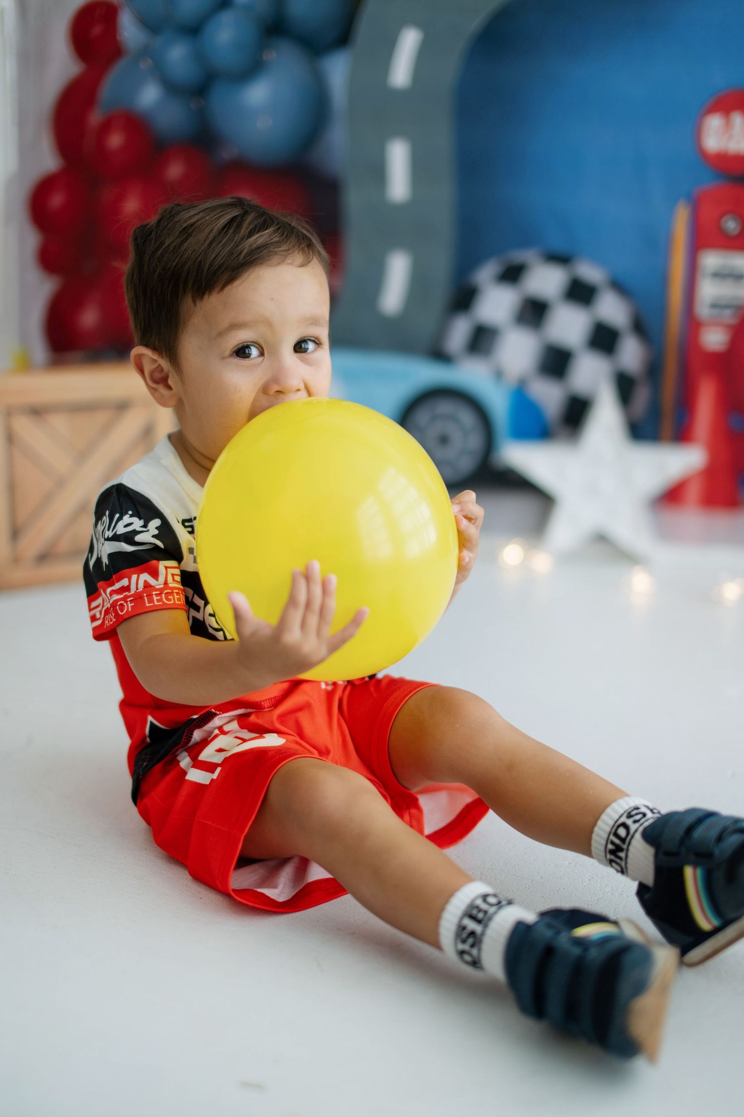 Toddler boy in red shorts sits on floor holding yellow balloon at a race car themed birthday party.