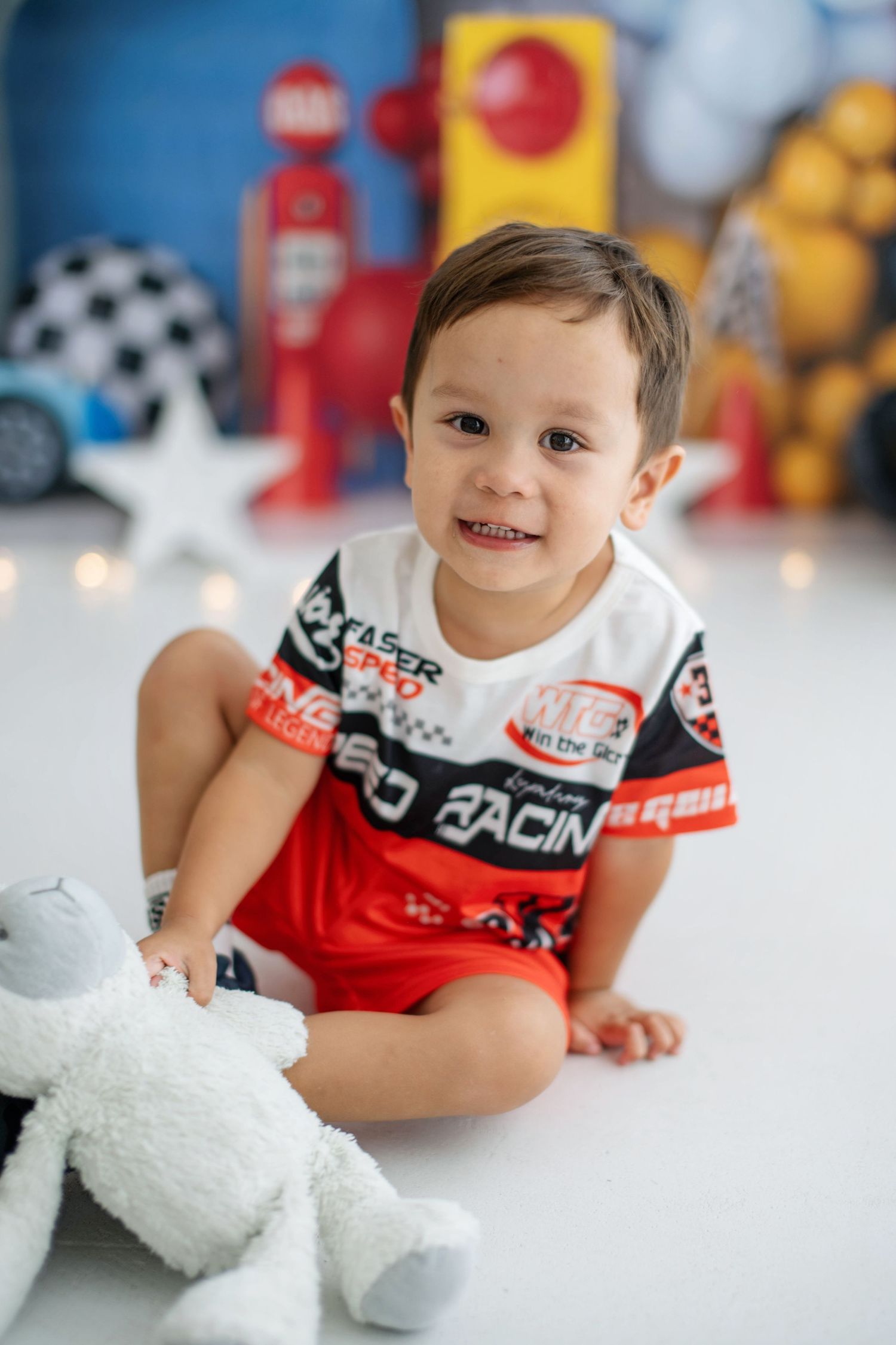 Smiling toddler boy in racing outfit sits on white floor with stuffed animal, surrounded by race car themed birthday decorations.