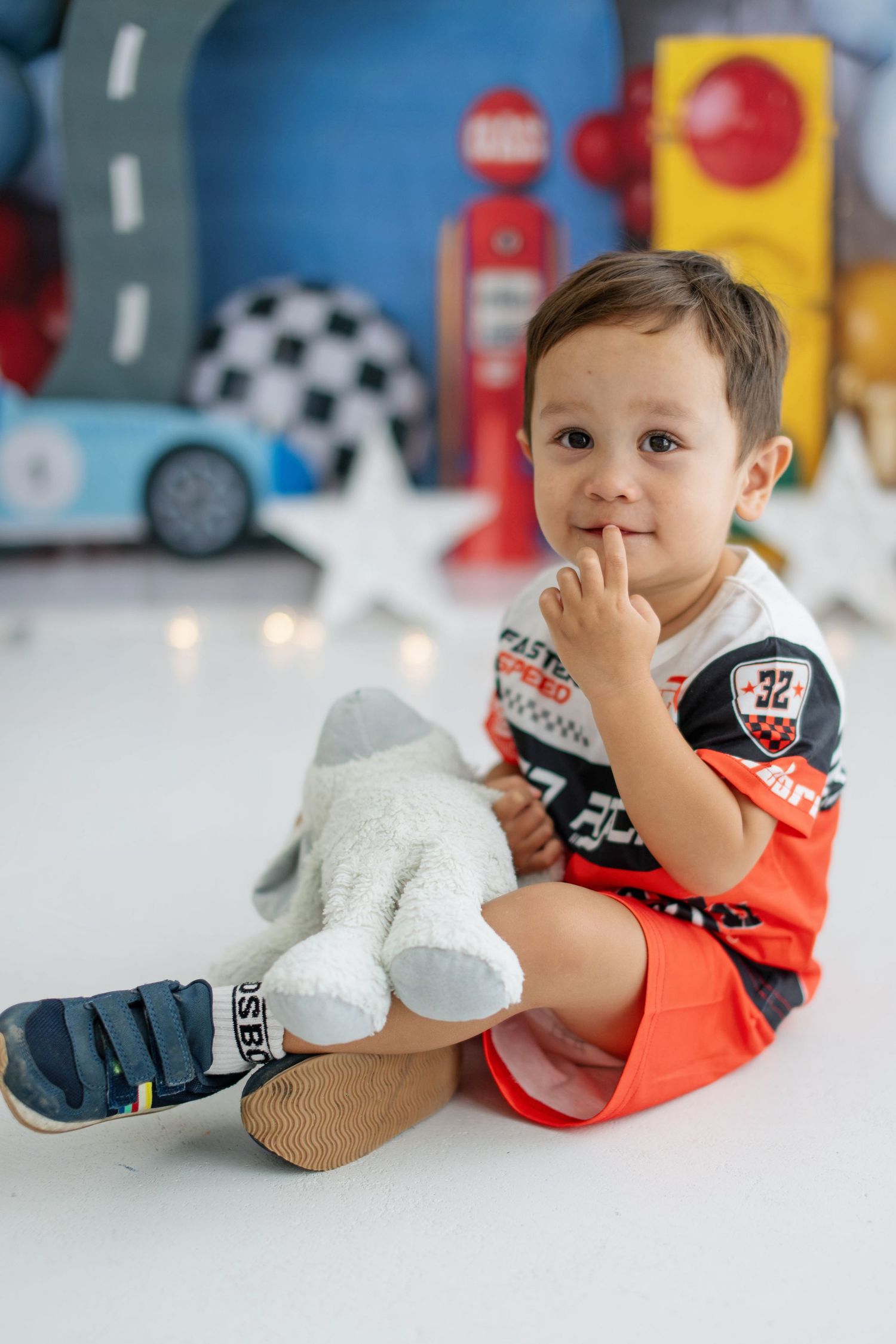 Cute toddler boy in racing outfit sits on floor holding stuffed elephant toy with race car themed backdrop.