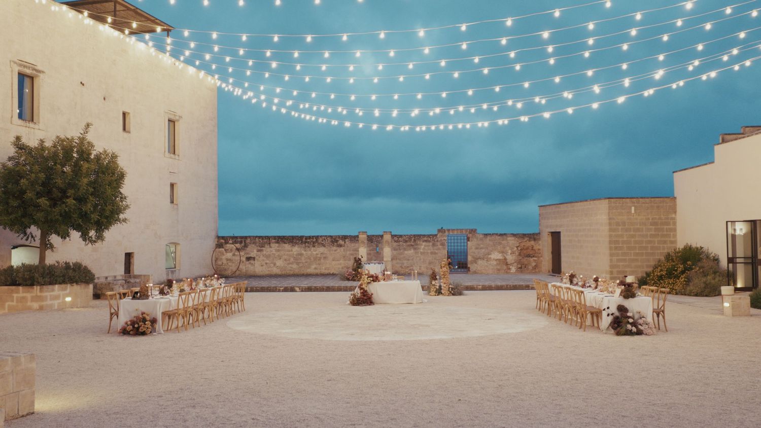 Elegant outdoor wedding venue with string lights canopy over ceremony chairs in a rustic stone courtyard at dusk.