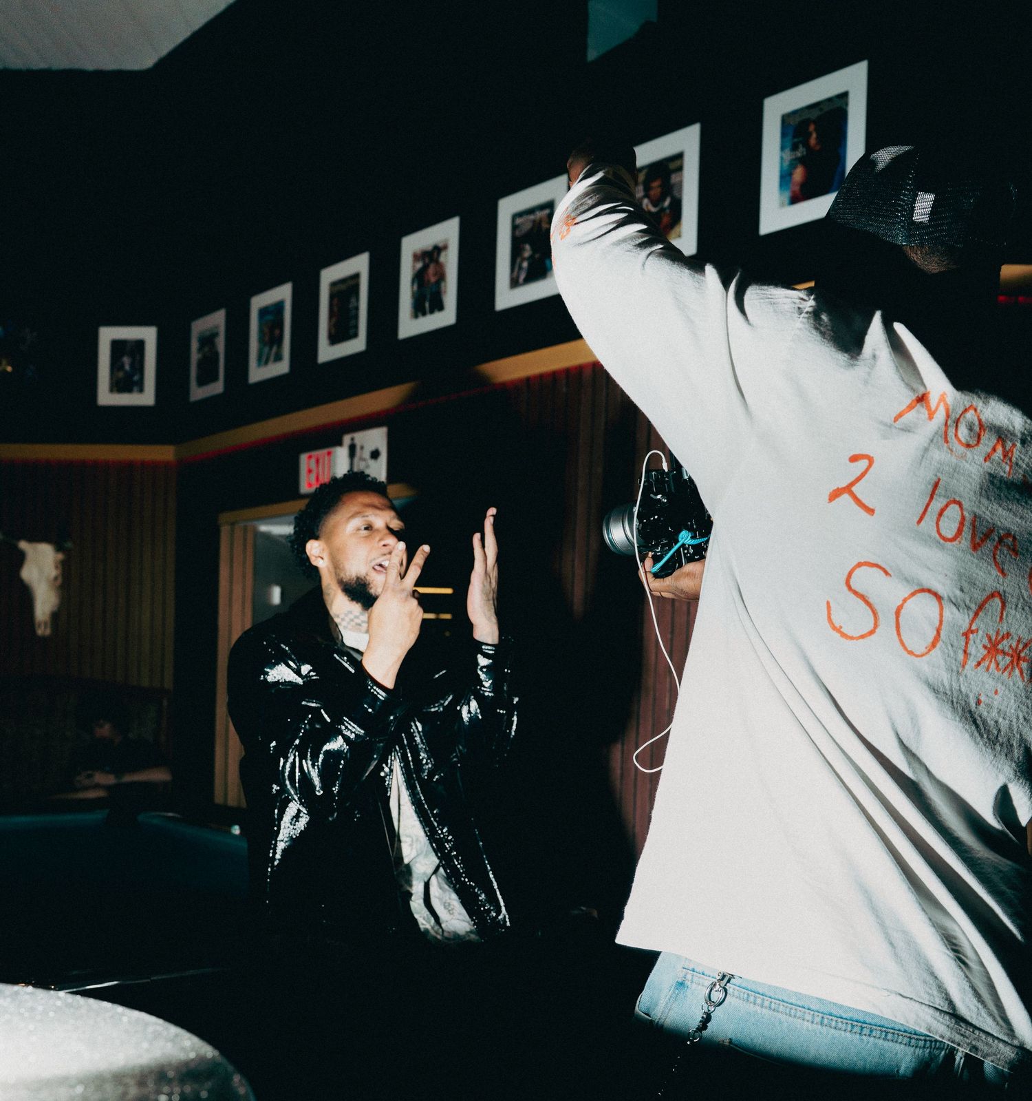Two men performing in a dimly lit room with framed photos on the wall, one in a shiny jacket, another in a white hoodie.
