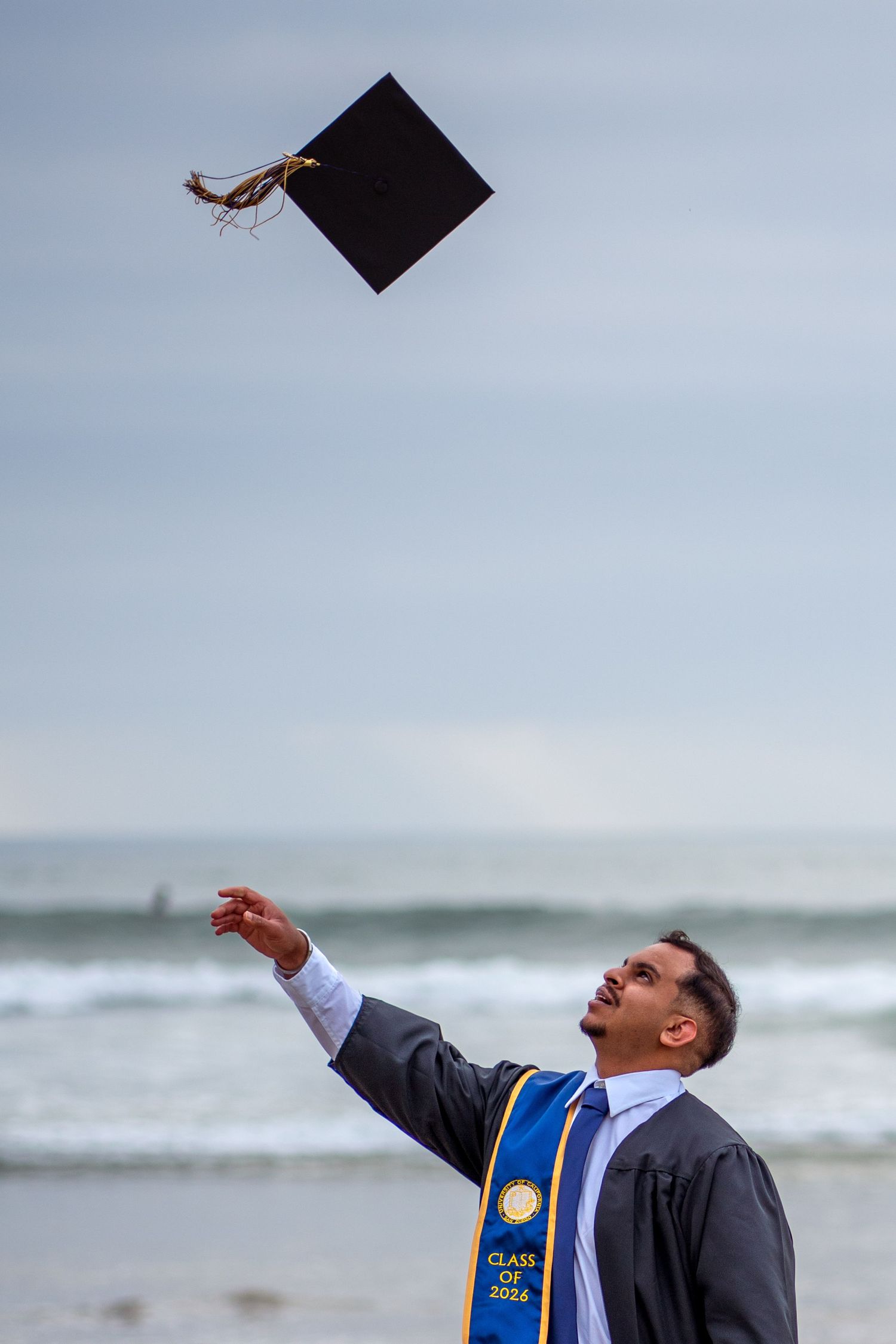 UCSD grad in grad gown and stole, tossing up his grad cap at a cloudy beach with waves in the background. La Jolla, CA
